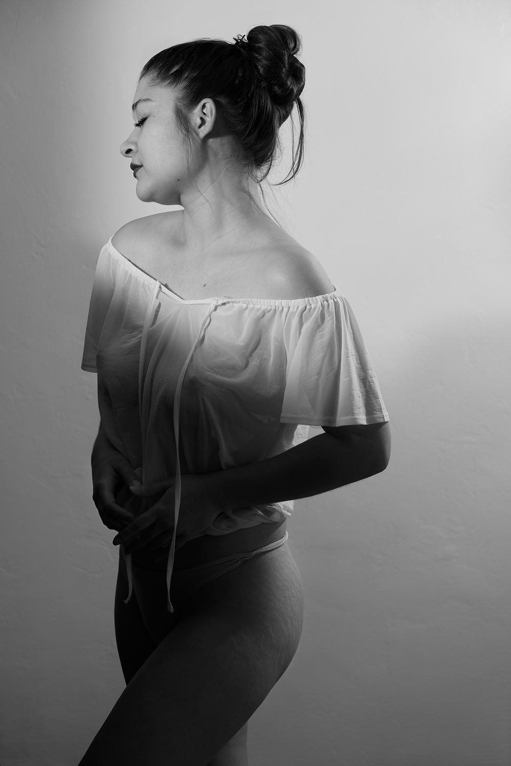 Black and white artistic portrait of a woman in an off-shoulder white top posing by a window.