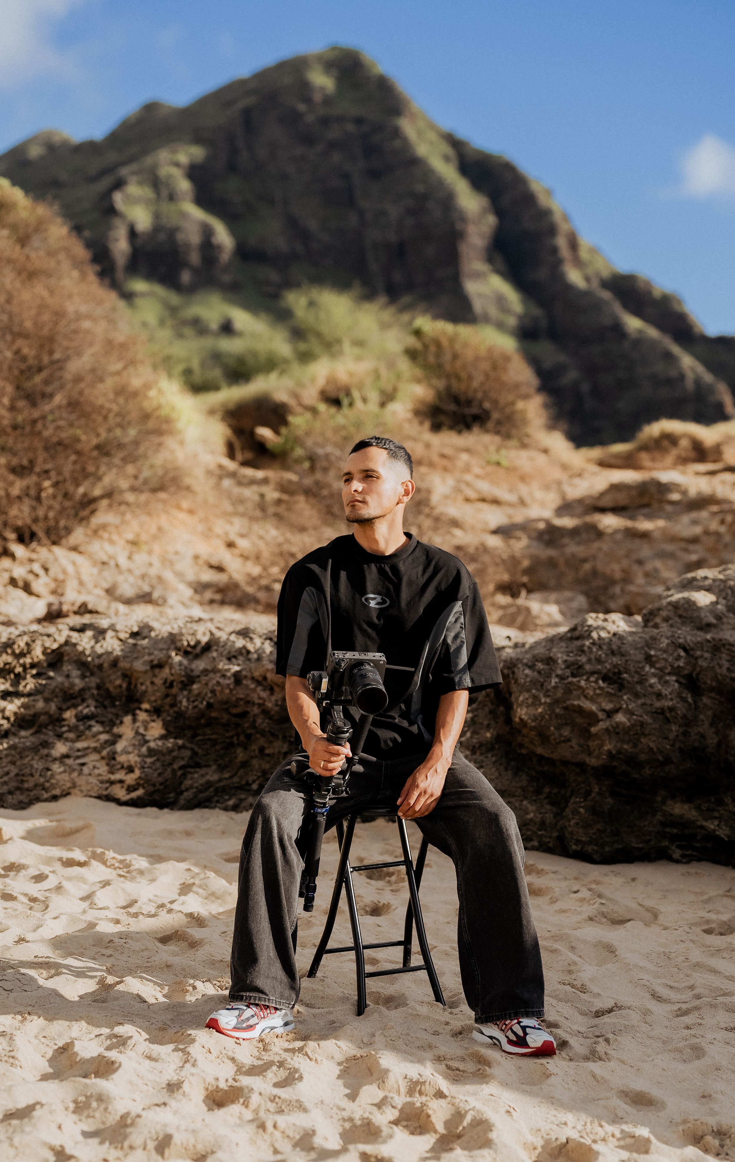 Photographer in black shirt and cap stands with professional camera equipment in outdoor natural setting.