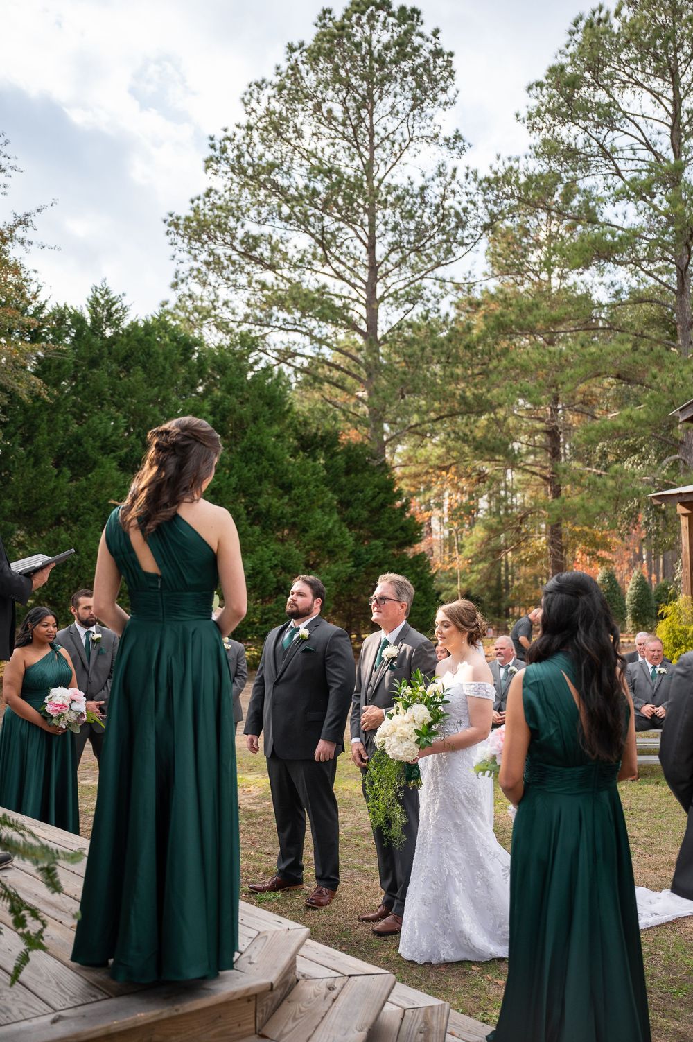 Wedding ceremony scene with bridesmaids in dark green dresses and guests outdoors.