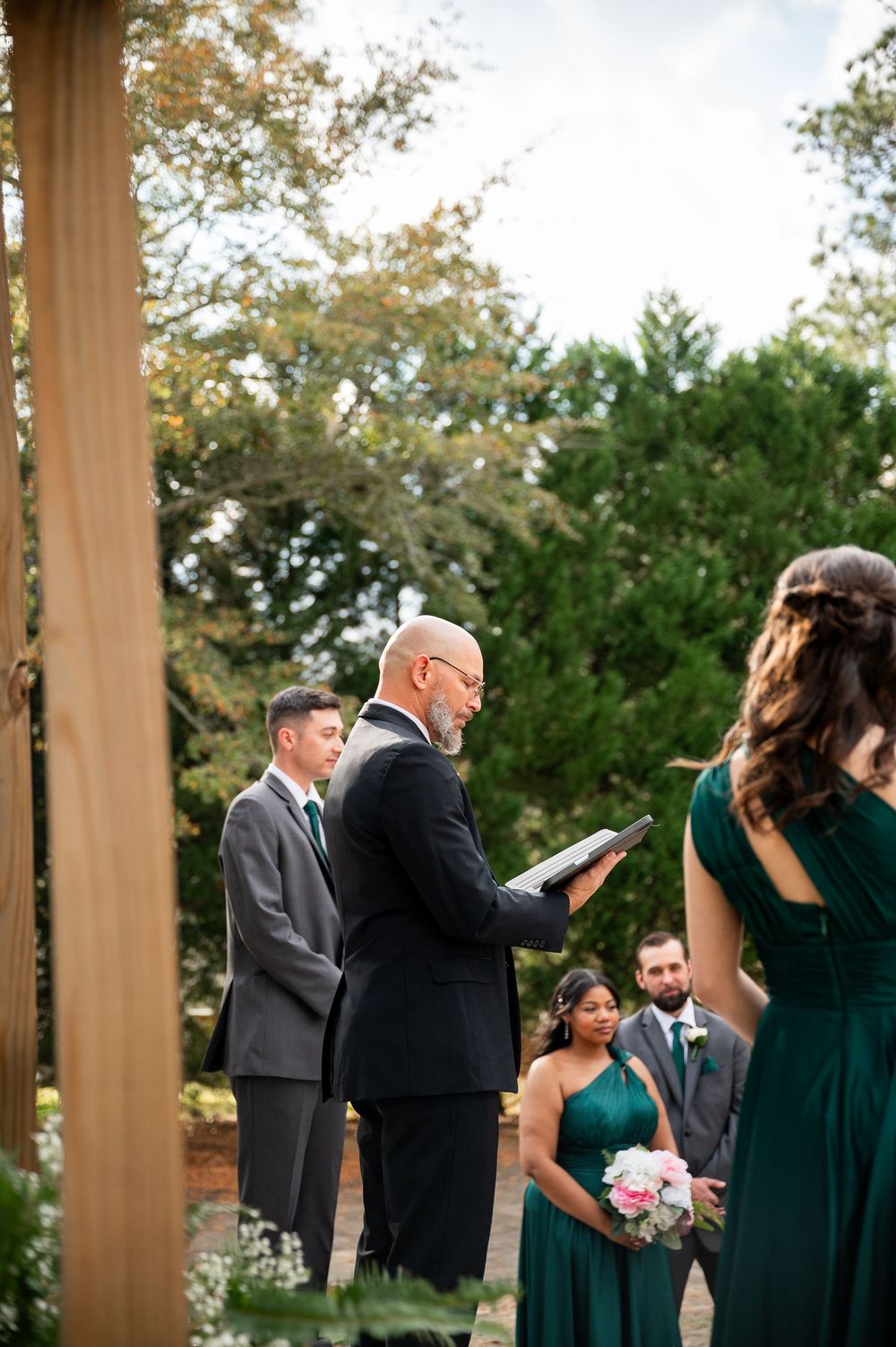 Wedding party members gather under a wooden pavilion during an outdoor ceremony.