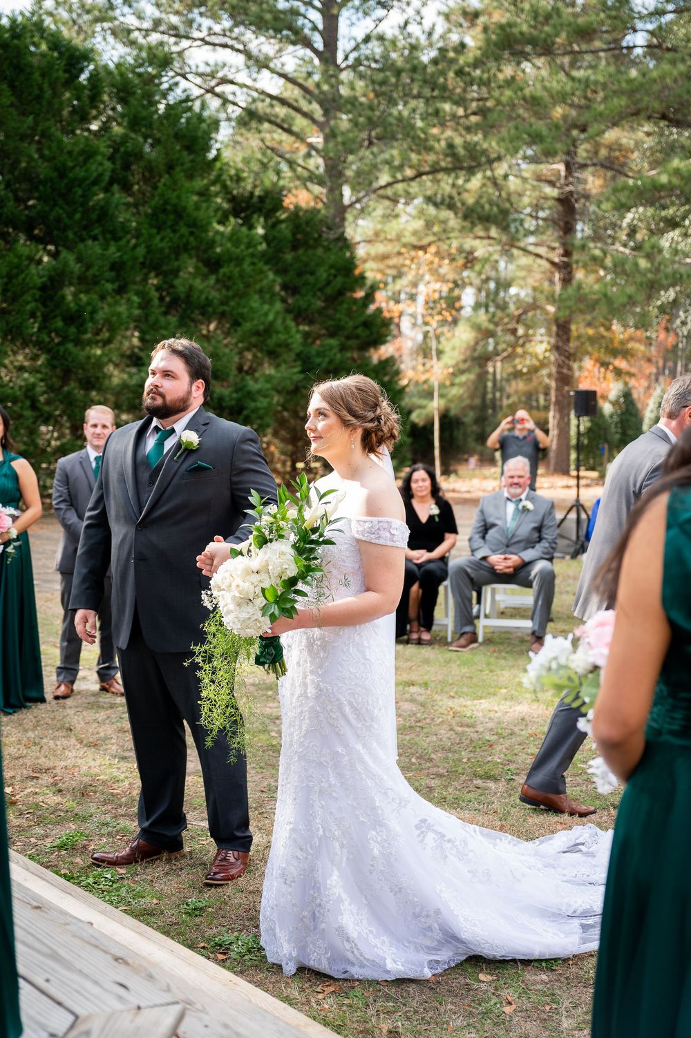 Bridal party in emerald green dresses and suits pose outdoors among pine trees.
