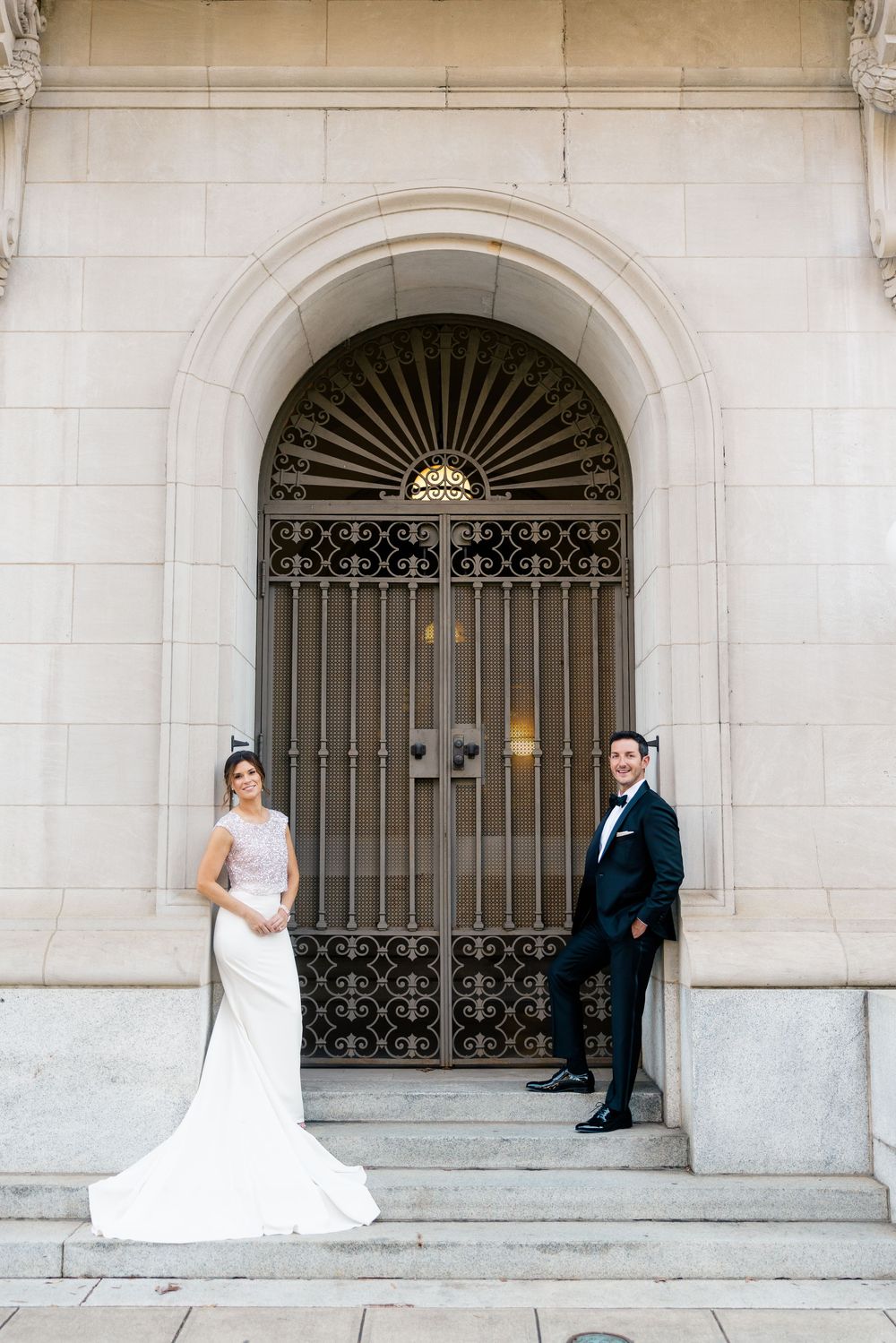 Elegant couple poses in formal attire on stone steps in front of ornate arched iron gate entrance.
