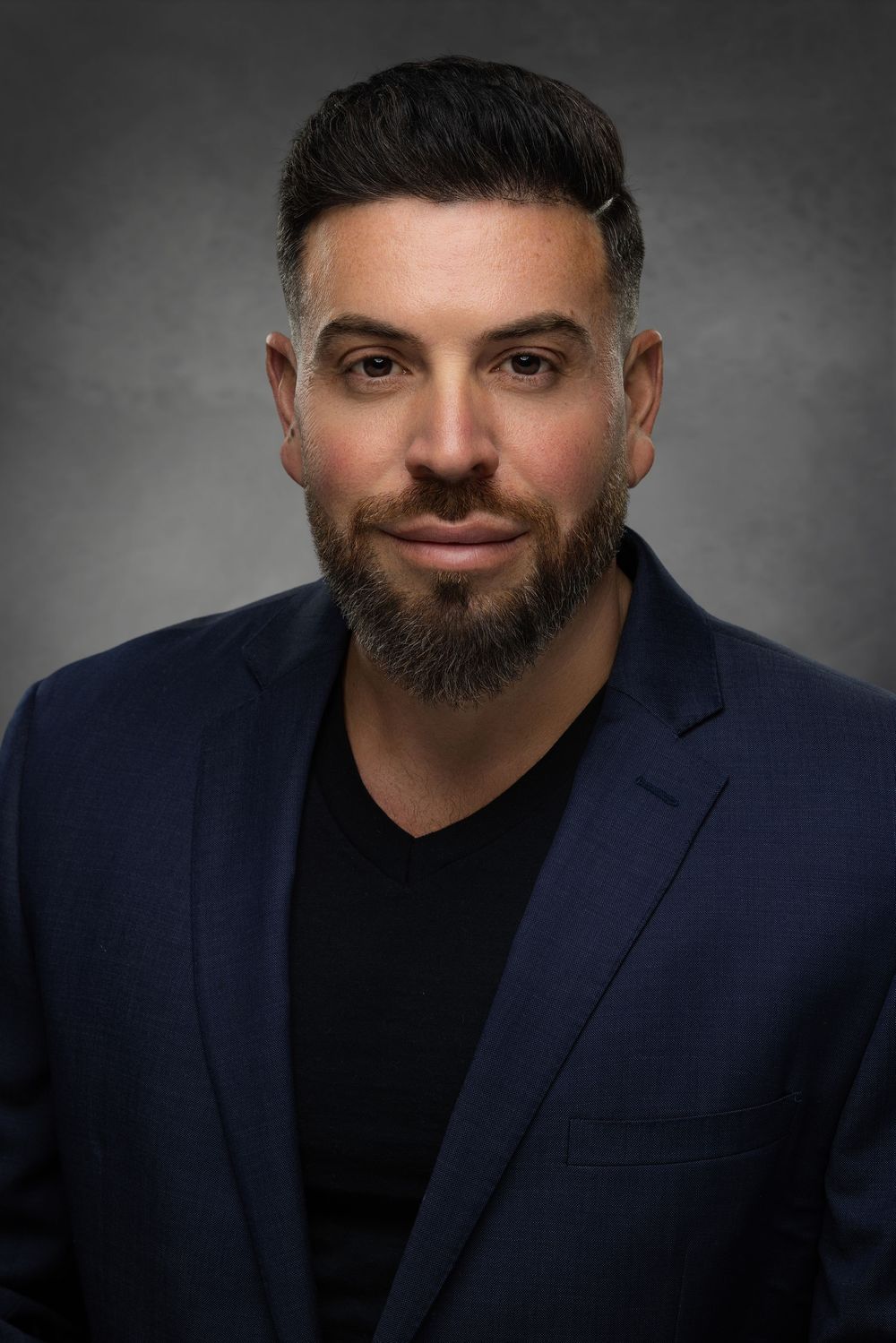 Professional studio portrait of a person in a navy blazer and black shirt against a dark gray background.