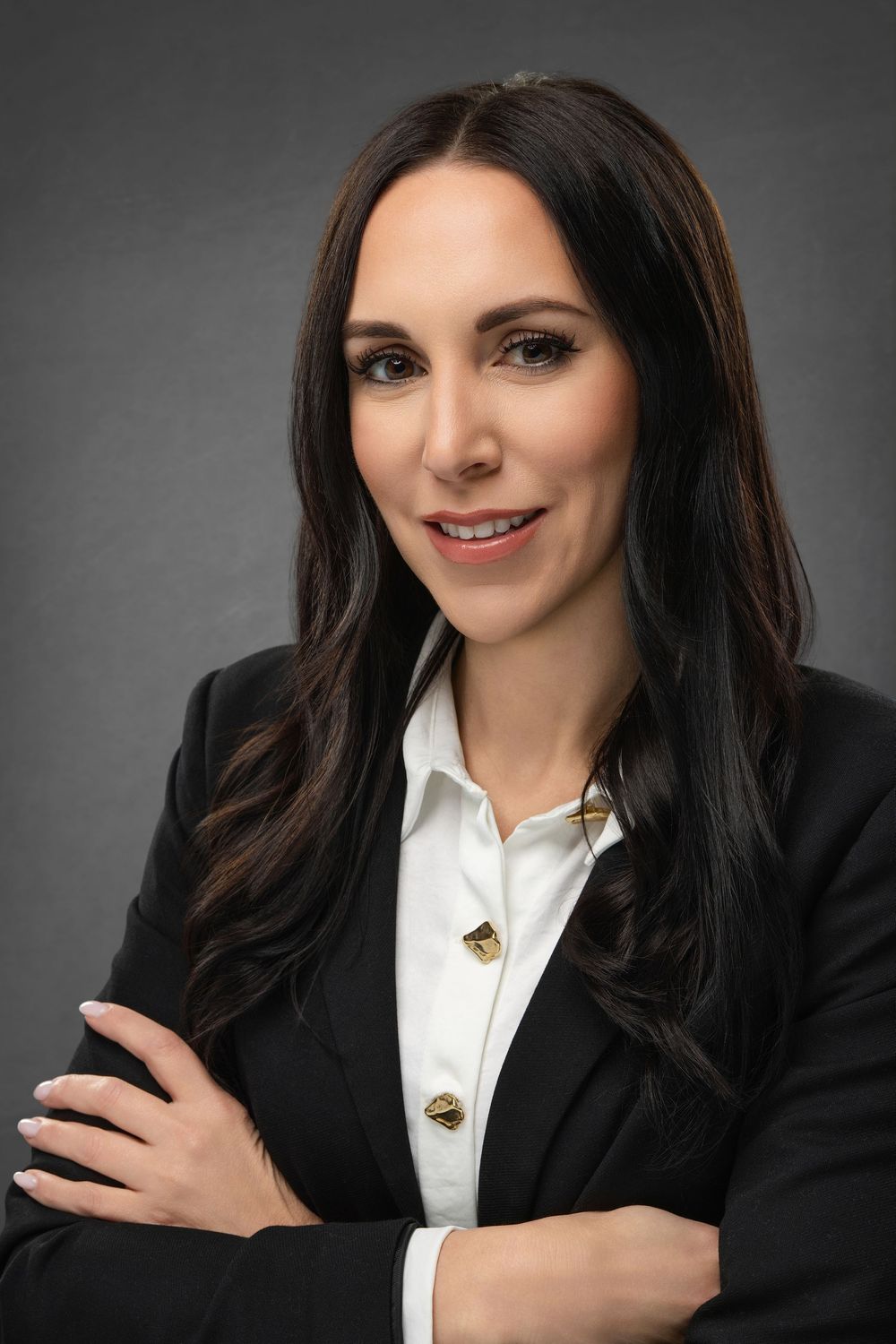 Professional business portrait showing a black blazer with white blouse and gold jewelry against a dark studio backdrop.