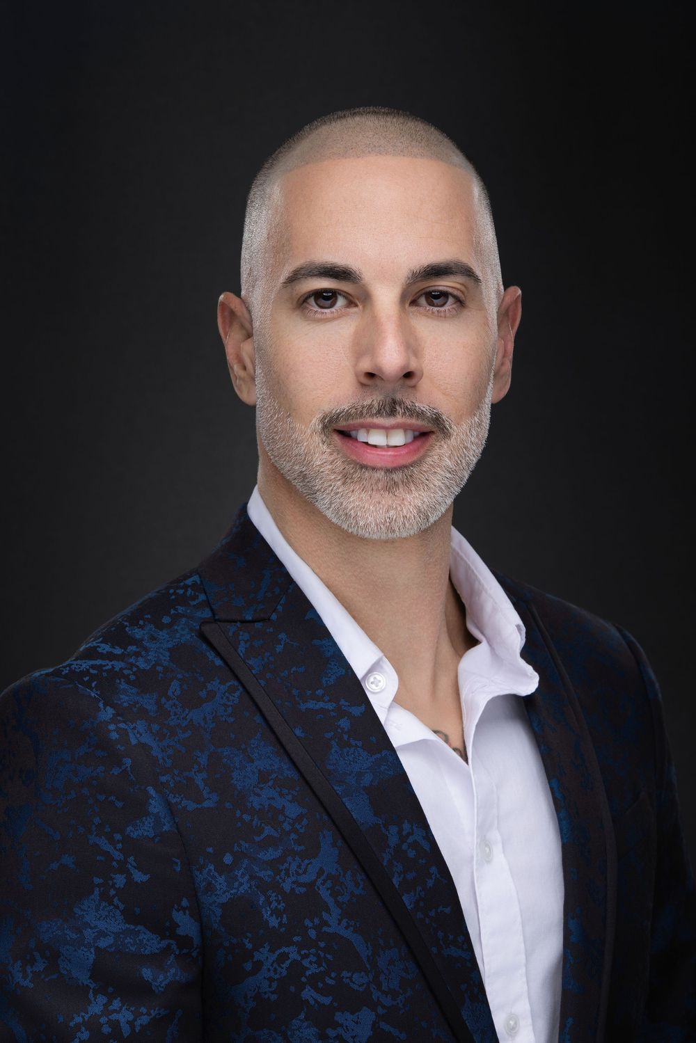 Professional headshot of a person wearing a blue patterned blazer and white dress shirt against a dark background.