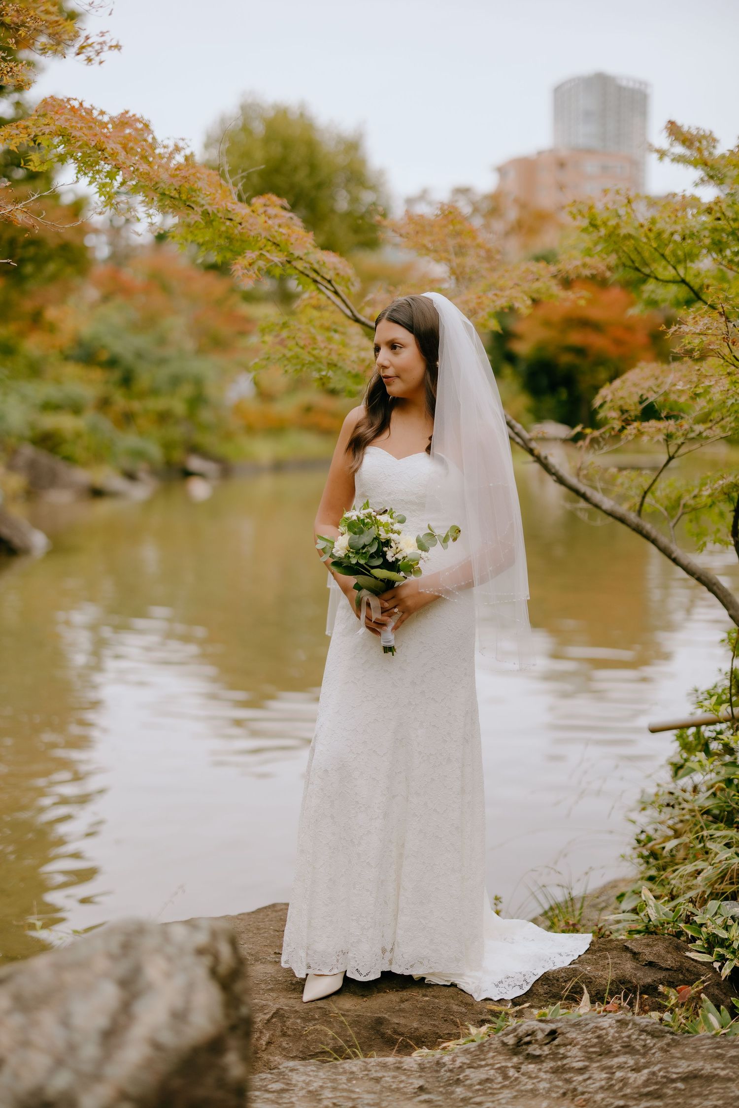 A bride in a flowing white dress and veil poses gracefully by a serene lake surrounded by autumn foliage.