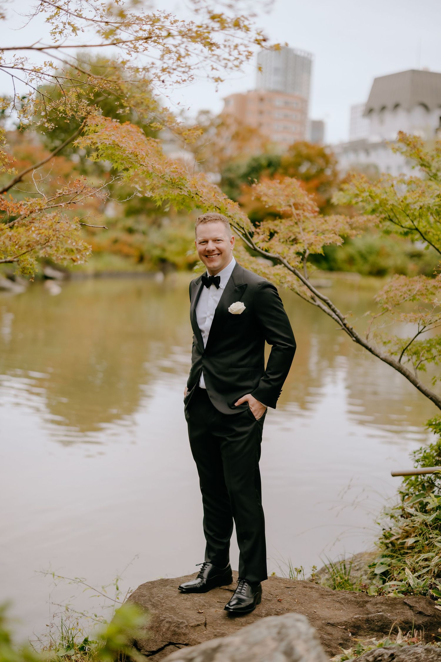 Groom in black tuxedo poses near autumn trees and lake during outdoor wedding photo session.