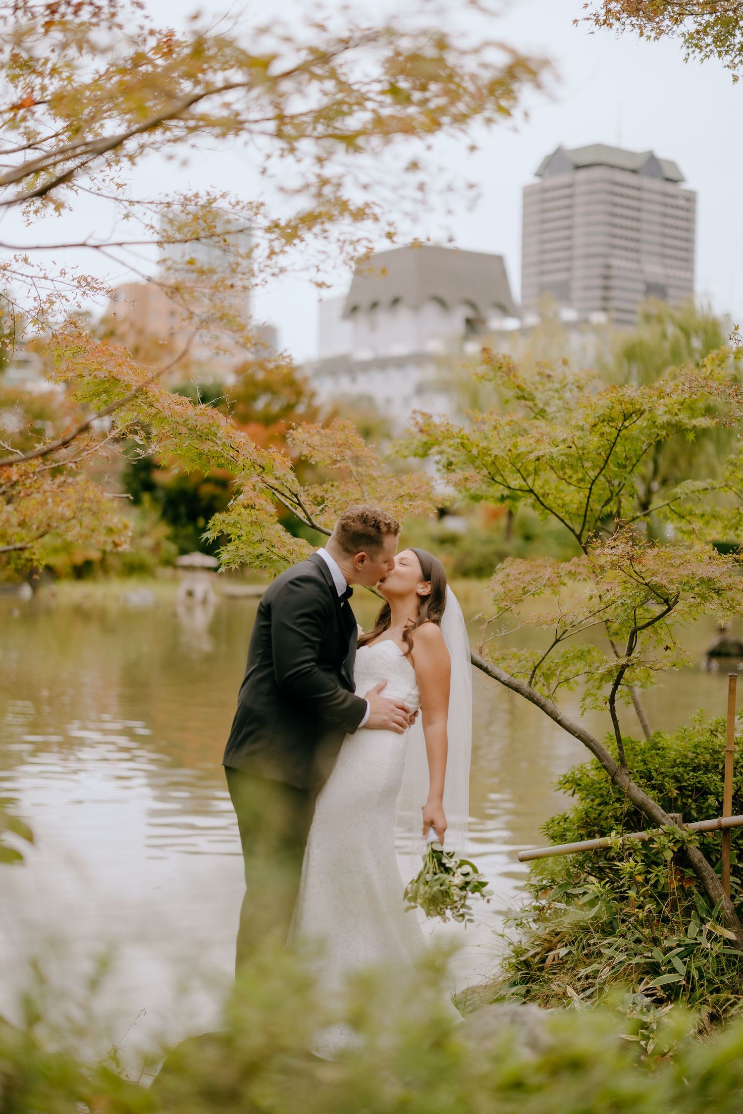 A romantic wedding couple shares a kiss by a serene pond in an autumn garden with fall foliage and city buildings in background.