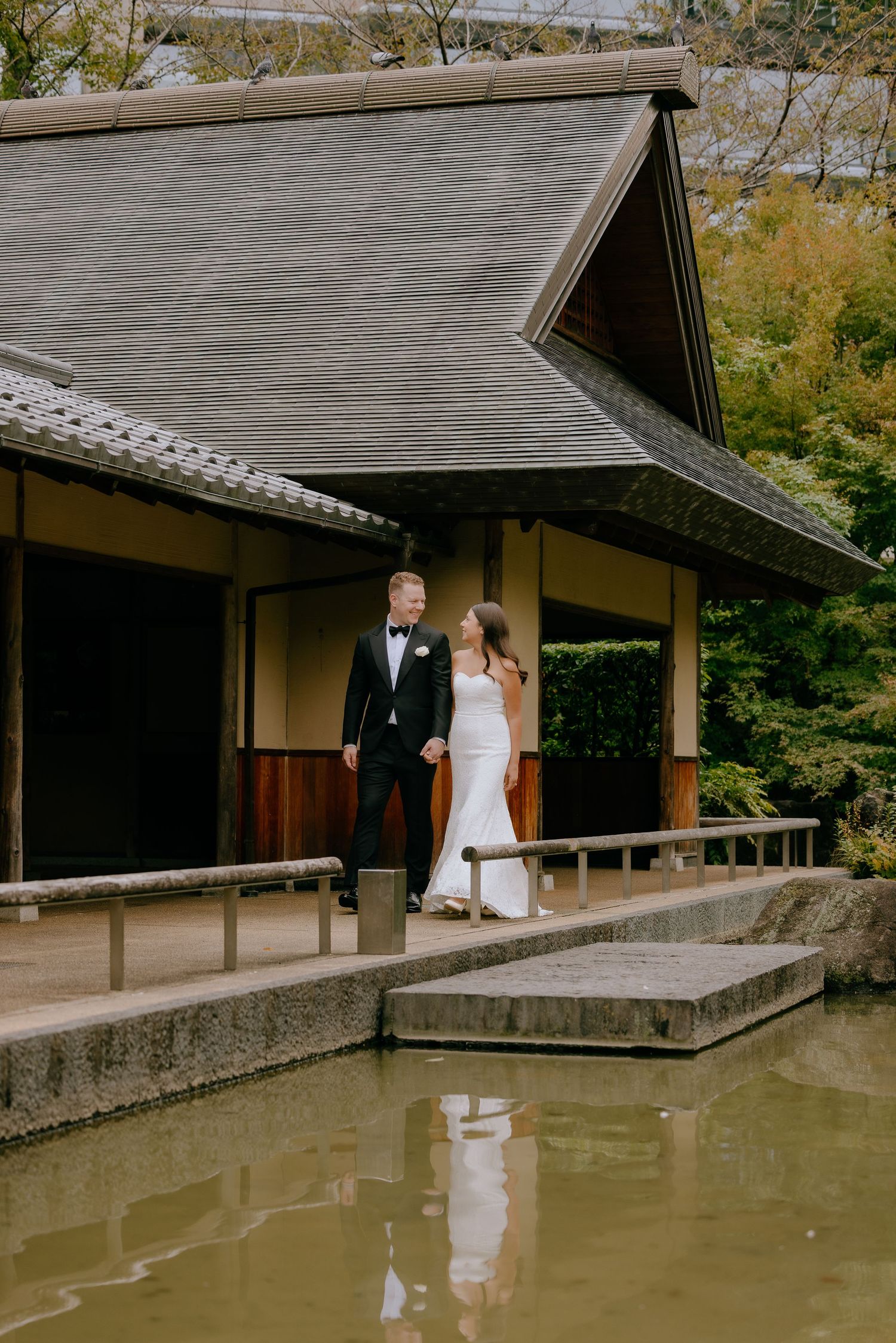 A couple in wedding attire stands by a tranquil pond at a traditional Japanese garden with wooden structures.