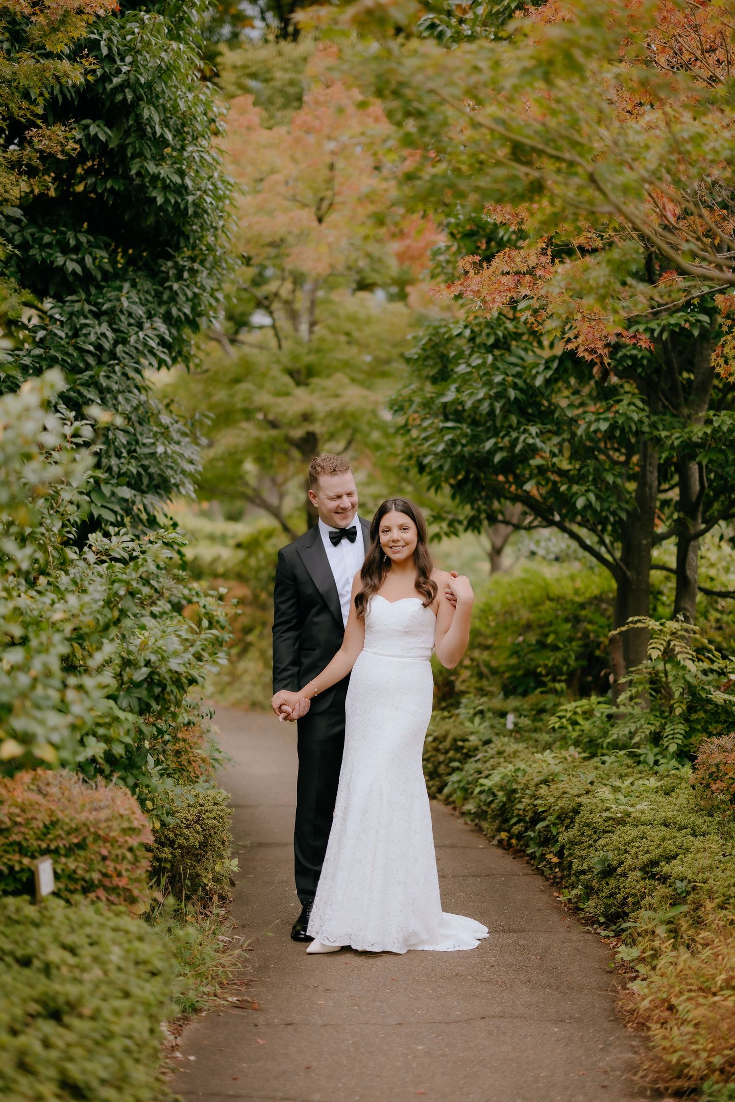 A couple walks together on a garden path surrounded by lush autumn foliage and greenery.