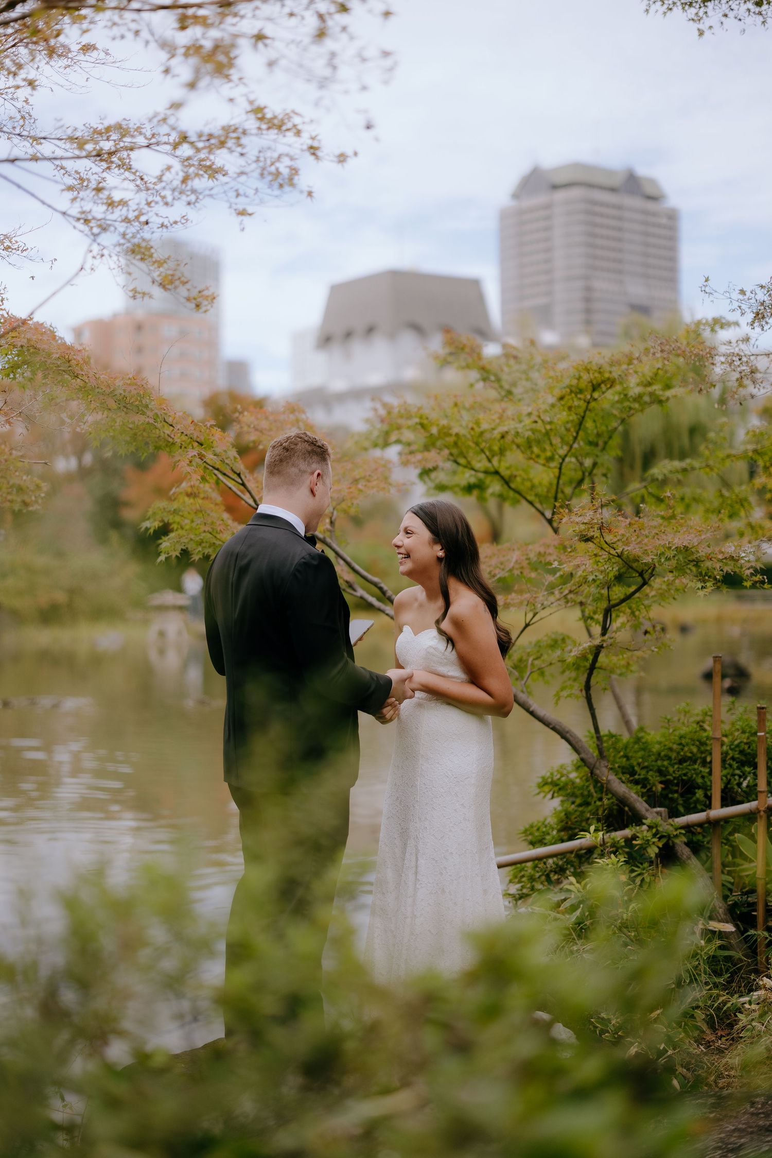A romantic couple shares intimate moments by a lake in an urban park during autumn with city buildings in the background.