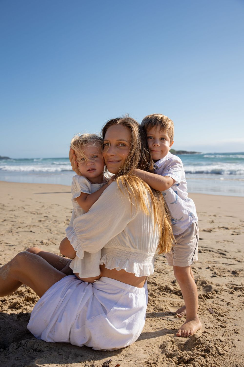 A family shares tender moments together while sitting on a sunny beach with ocean waves in the background.