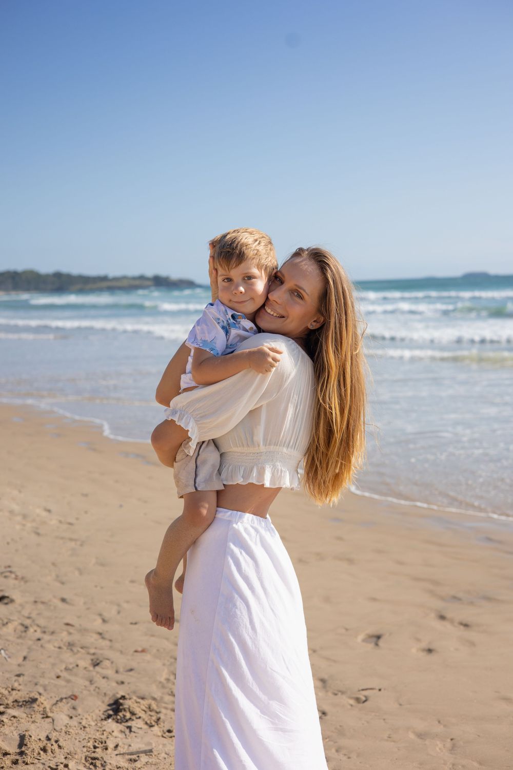 A parent and child share tender moments together while walking along a sunny beach with crashing waves.