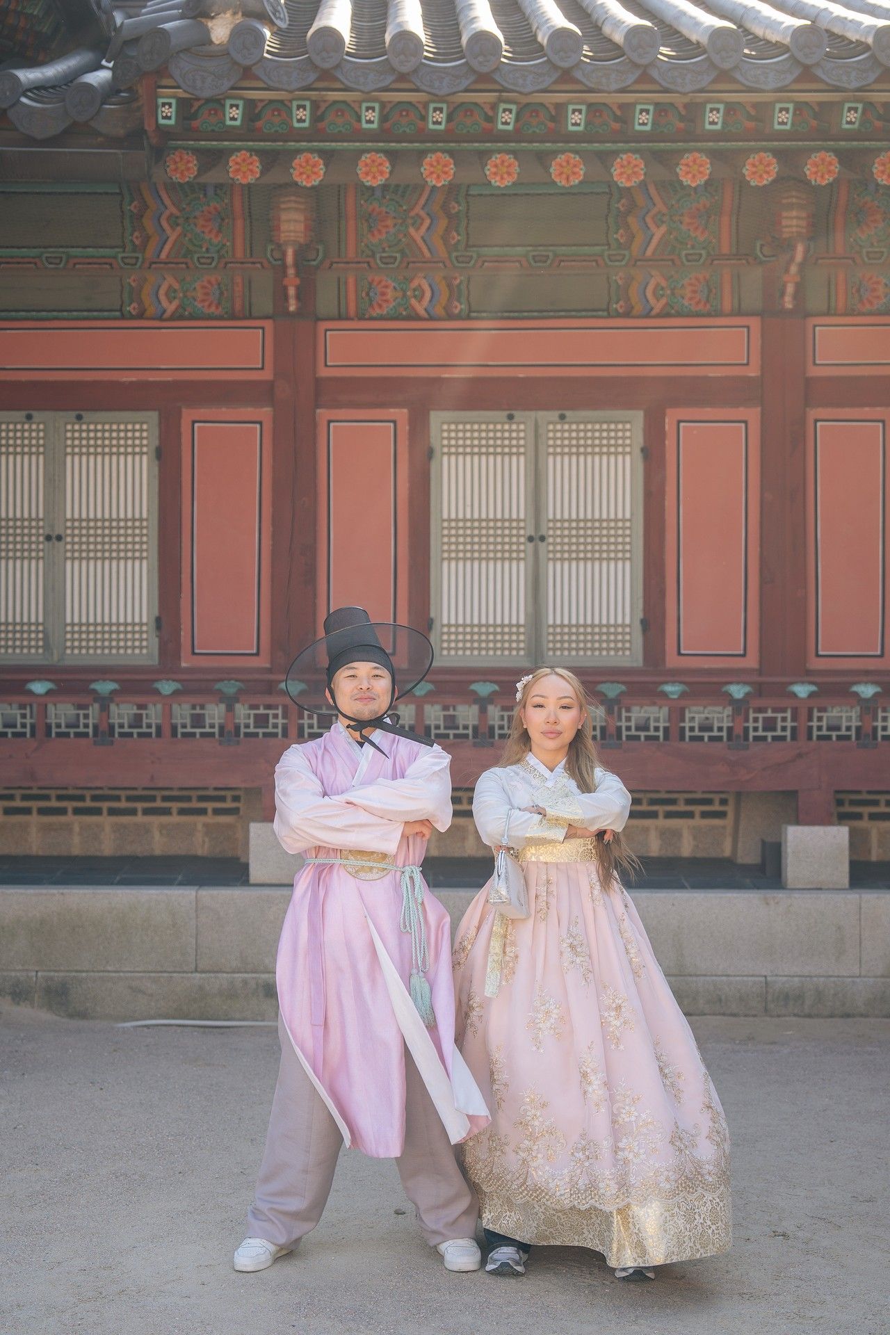 Two people wearing traditional Korean hanbok outfits pose together in front of a historic palace building in Seoul.