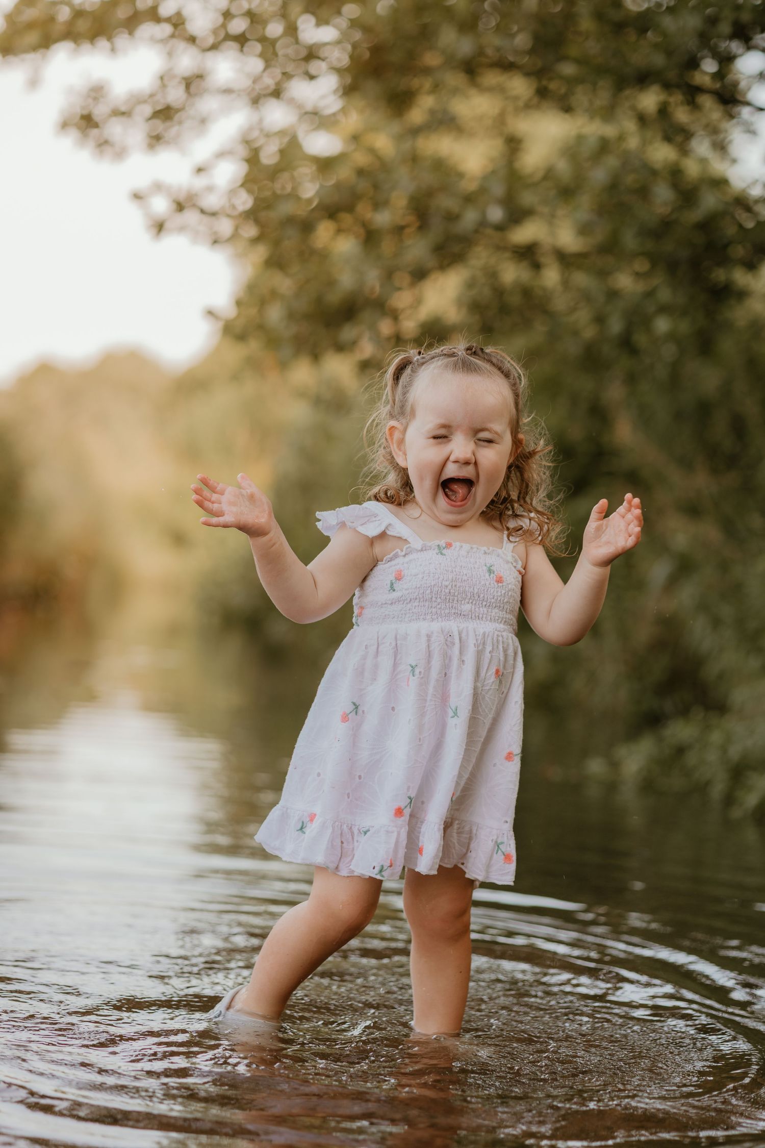 A small child wearing a white dress plays joyfully in shallow water during golden hour.