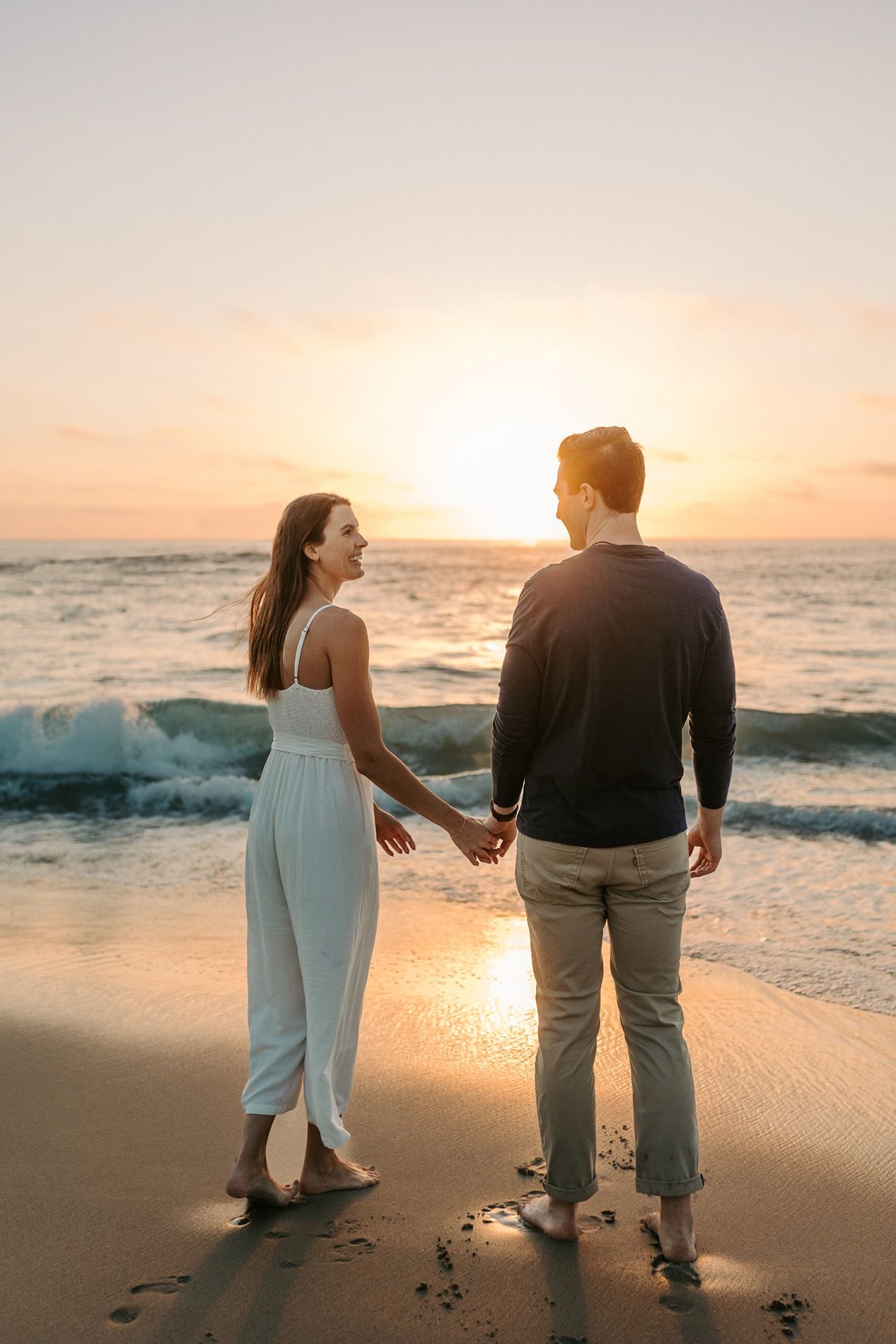 Couple walking along beach at sunset with waves crashing and footprints in sand creating romantic silhouettes at Windansea Beach.