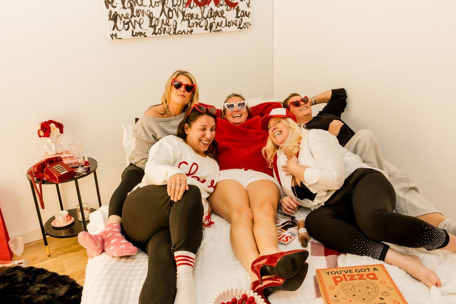 A group of besties laying in bed laughing while on set of a messy red, black, and white bedroom during a photoshoot.