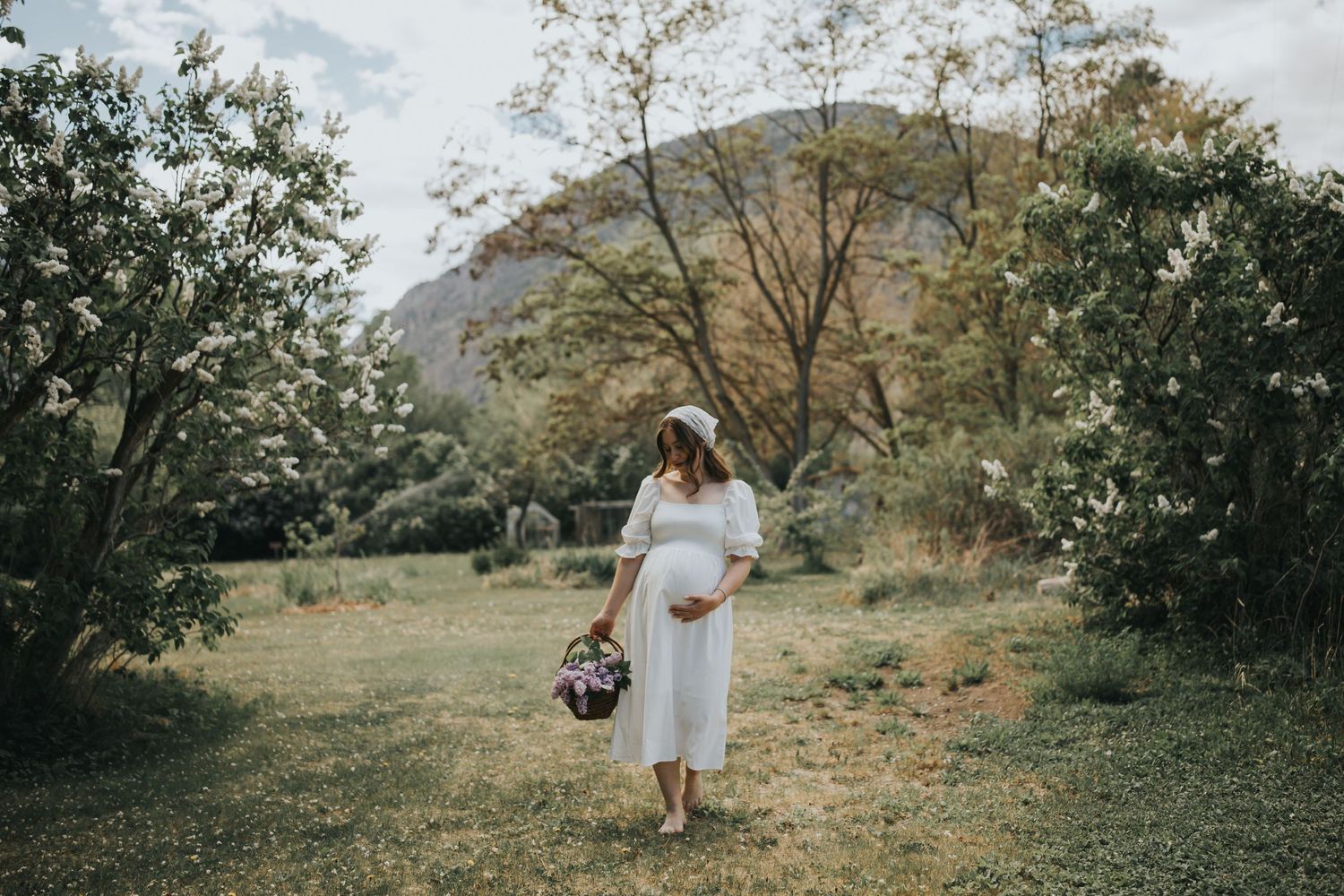 Outdoor maternity photo near Penticton BC featuring pregnant woman in a white dress and cowboy boots standing in a rustic fence and field.