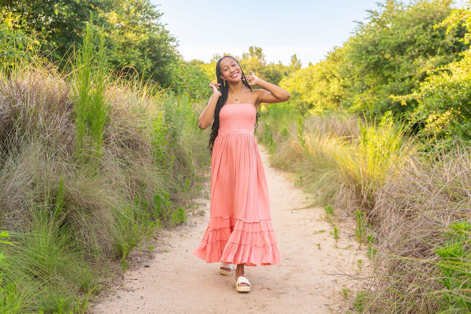A person in a flowing coral dress walks along a winding nature path surrounded by tall grasses and greenery in summer.