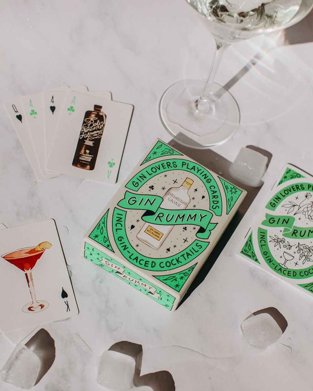 Playing cards featuring cocktail designs spread out with a rummy deck box and martini glass on white surface.