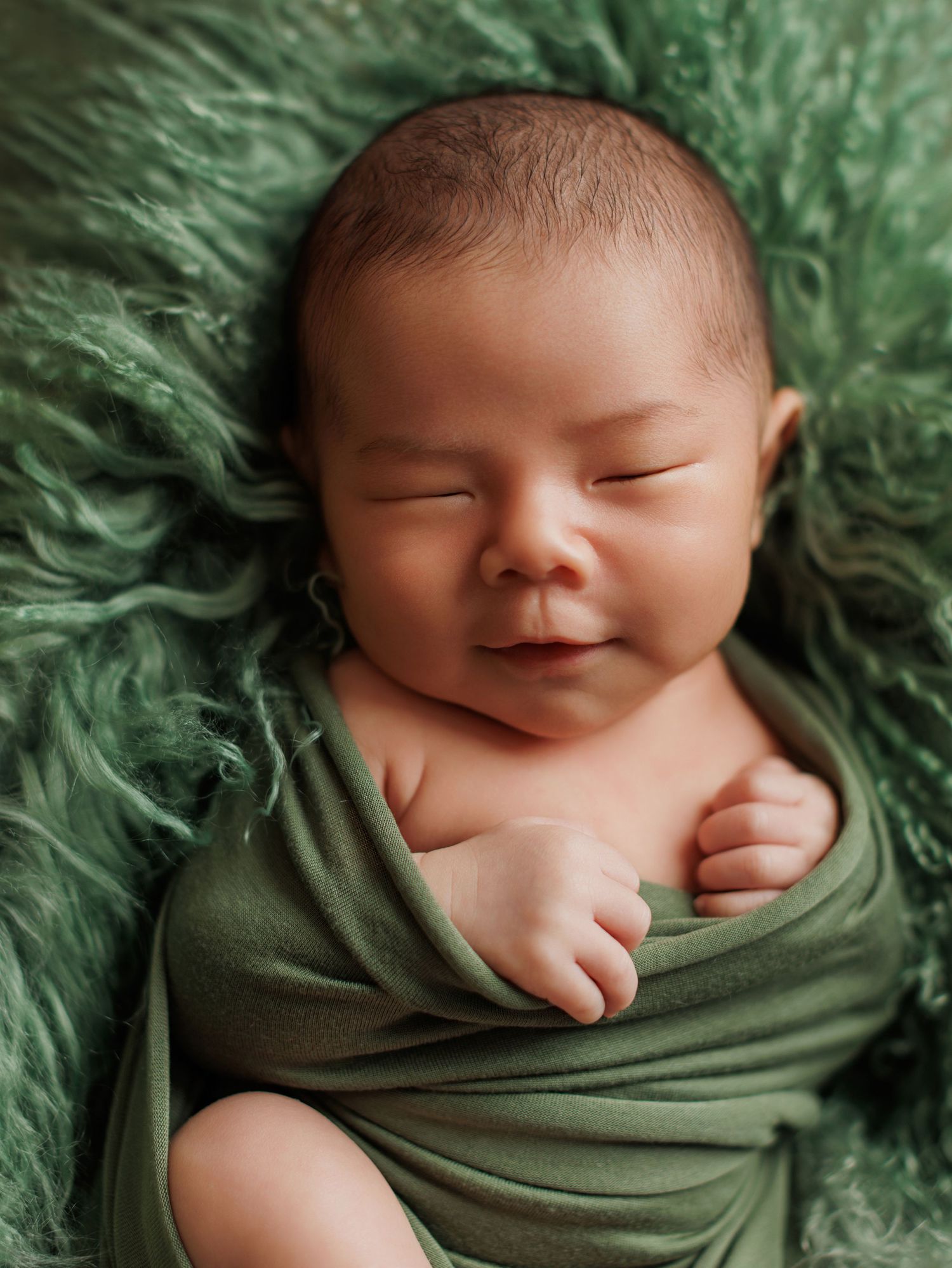 A peaceful sleeping newborn swaddled in olive green fabric against a fluffy green textured background.