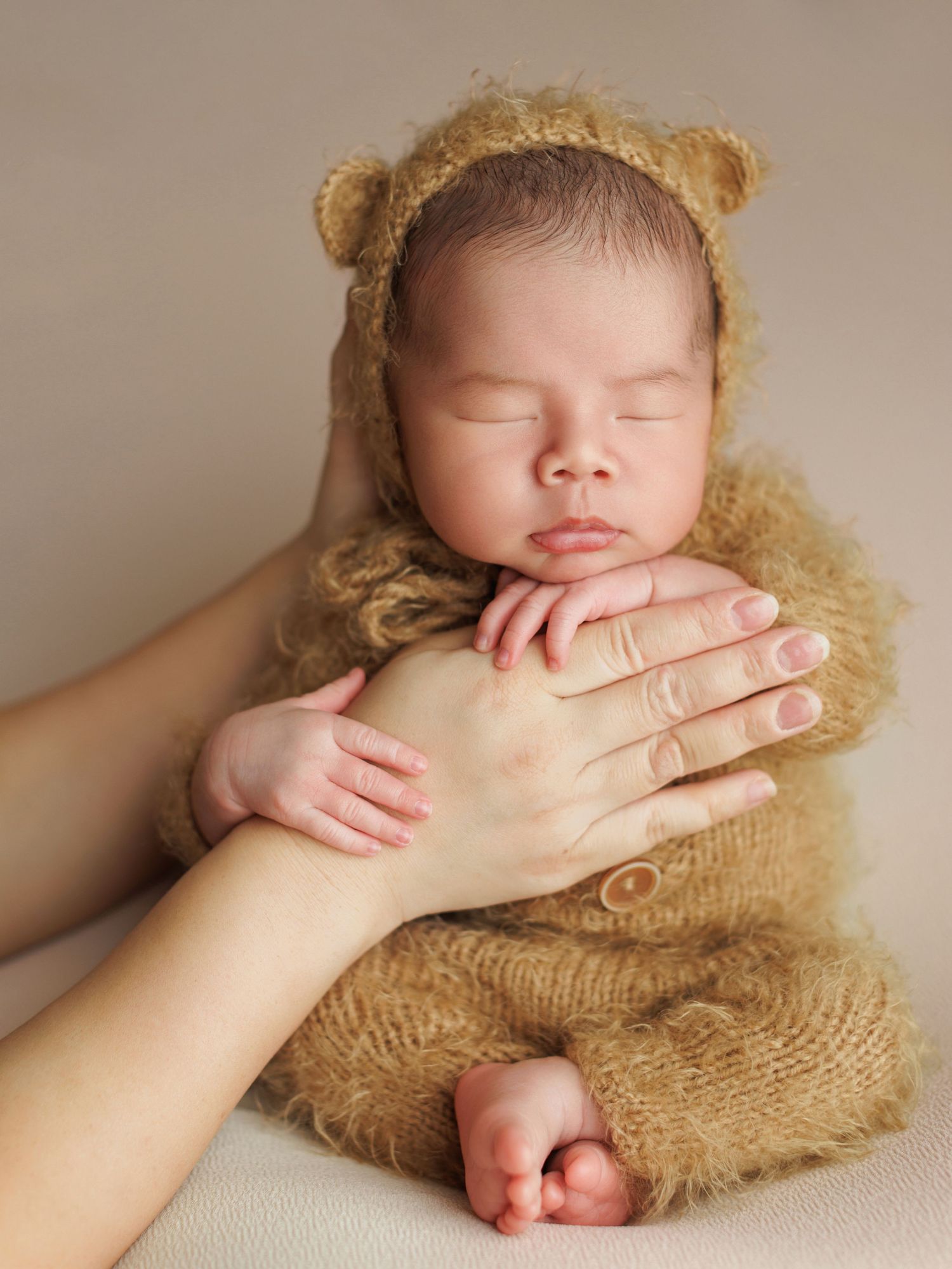 A sleeping newborn wrapped in a soft brown knit blanket with a hand gently covering their face.