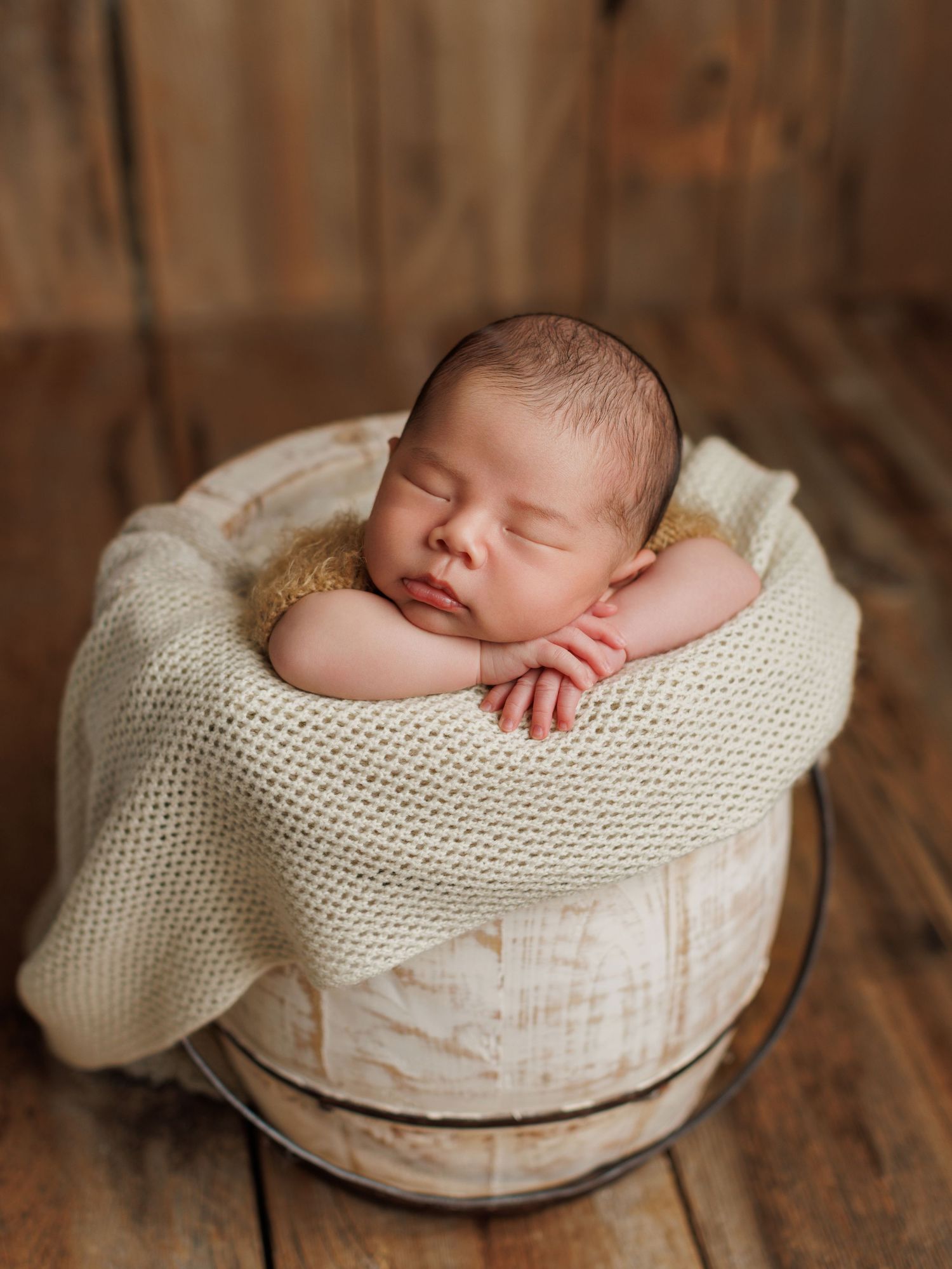 A sleeping newborn baby nestled in a cream-colored knit blanket inside a rustic wooden bowl on a textured surface.