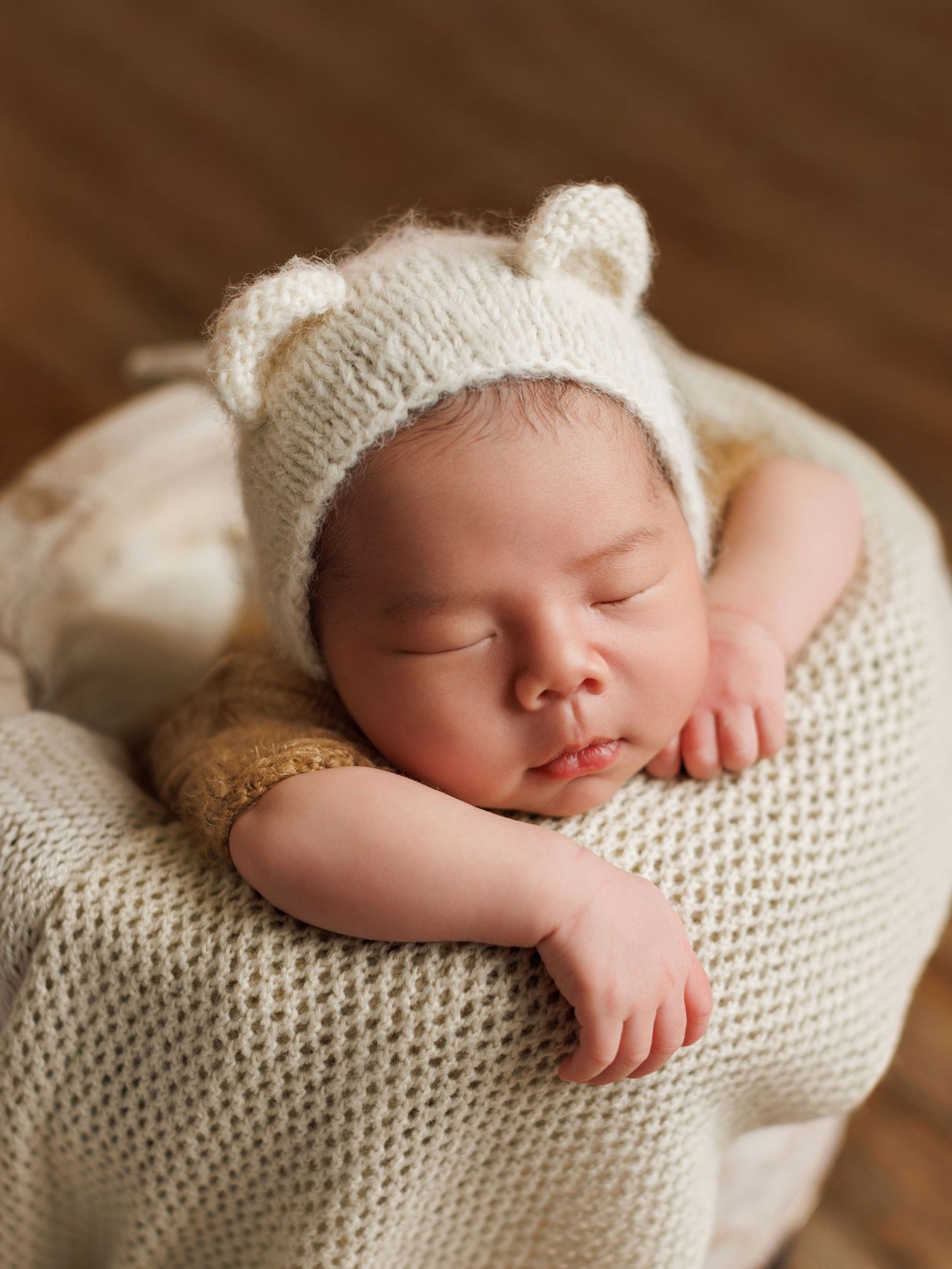 Sleeping newborn wearing white knit hat rests peacefully on cream-colored knitted blanket in warm natural lighting.
