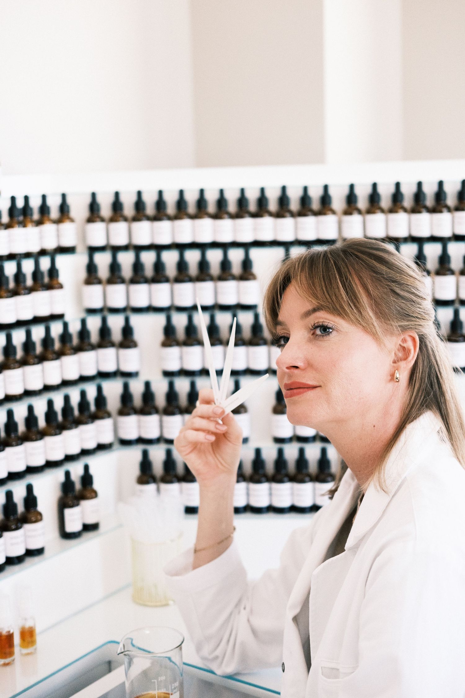 Perfumer in white lab coat examining fragrance samples in front of wall of essential oil bottles in laboratory.