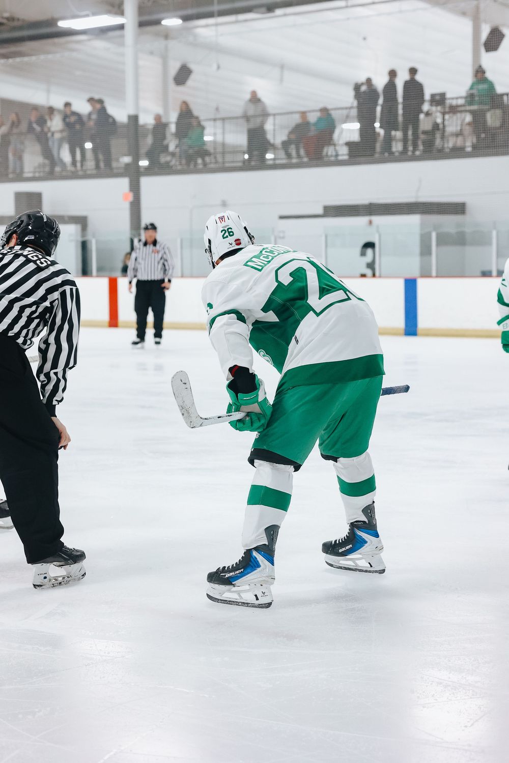 Hockey players and referee on ice during an indoor game with spectators in background.