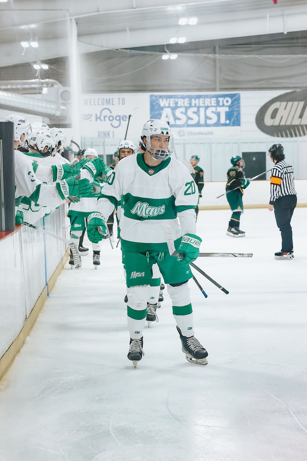 Hockey player in white and green uniform skating on ice during game with team banner visible.