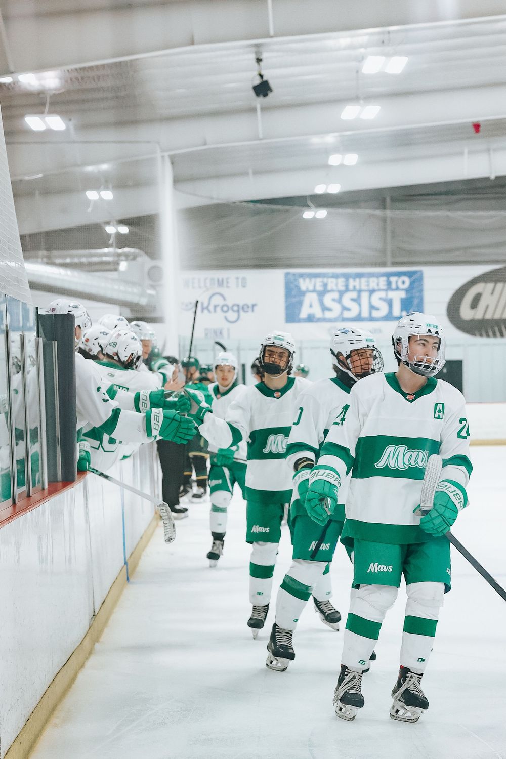 Hockey team in green and white uniforms skating in line along boards during game.