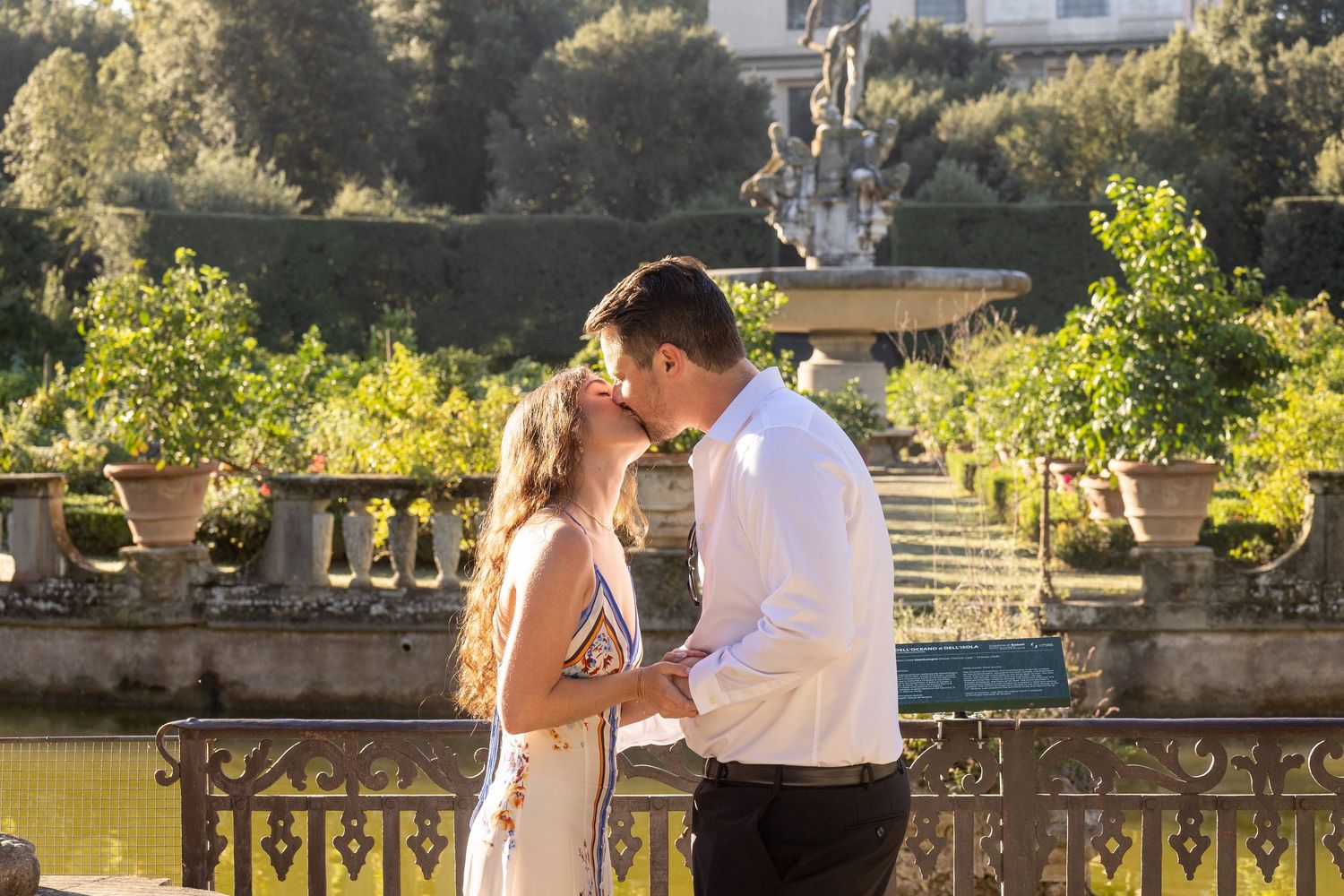 Outdoor wedding ceremony at sunset with guests gathered in a garden setting near stone columns and potted plants.
