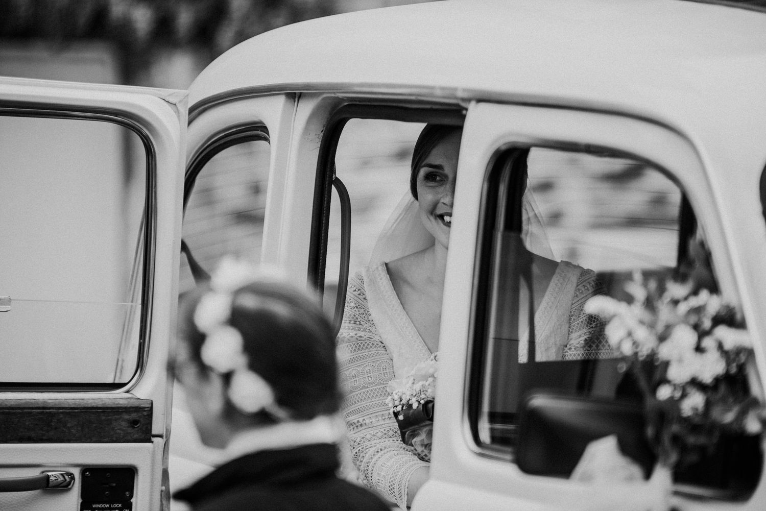 Mariée souriante dans une voiture ancienne le jour de son mariage, photo reportage en noir et blanc.