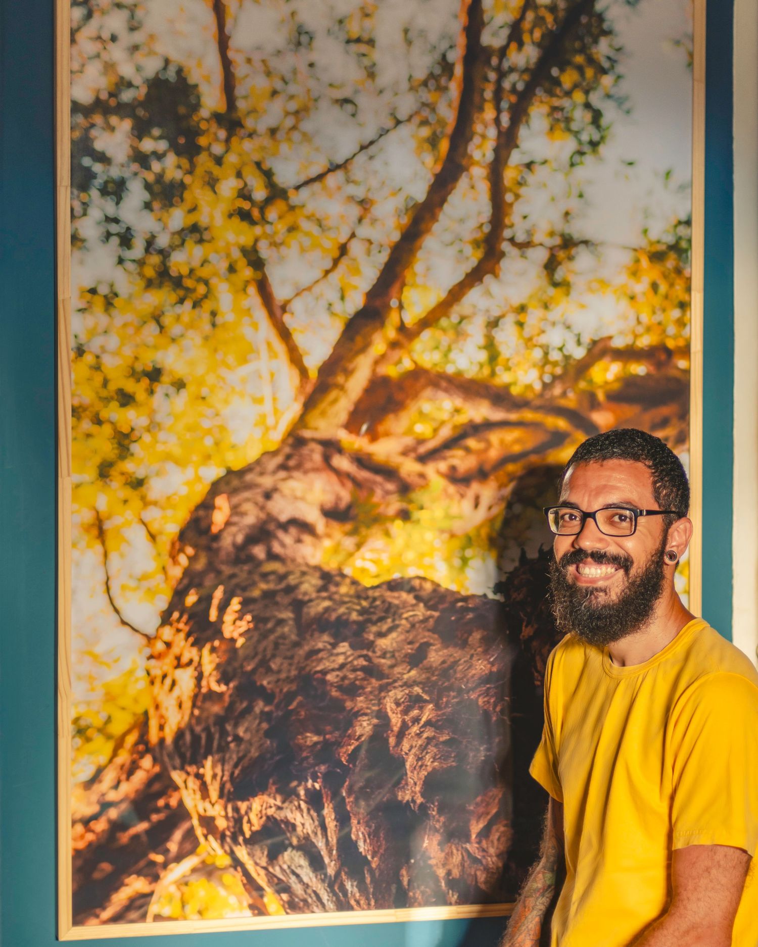 Fotógrafo Paulo Félix sorrindo ao lado de um quadro decorativo de arte. Contato para retratos sociais, corporativos e venda de quadros.