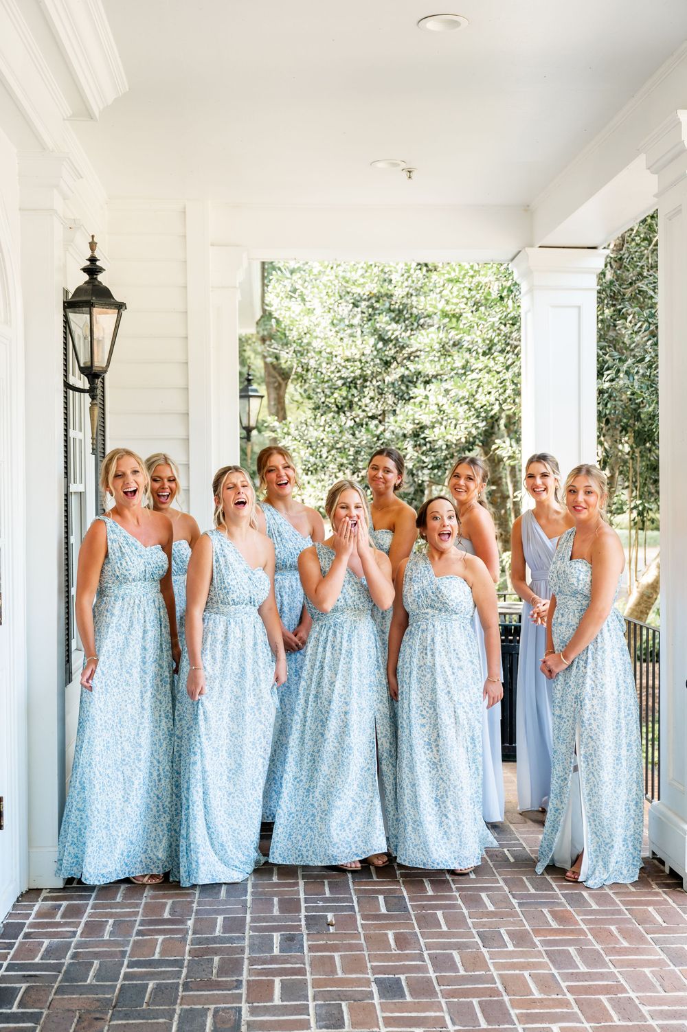 Bridesmaids in matching light blue floral dresses pose together on a covered porch with white columns.
