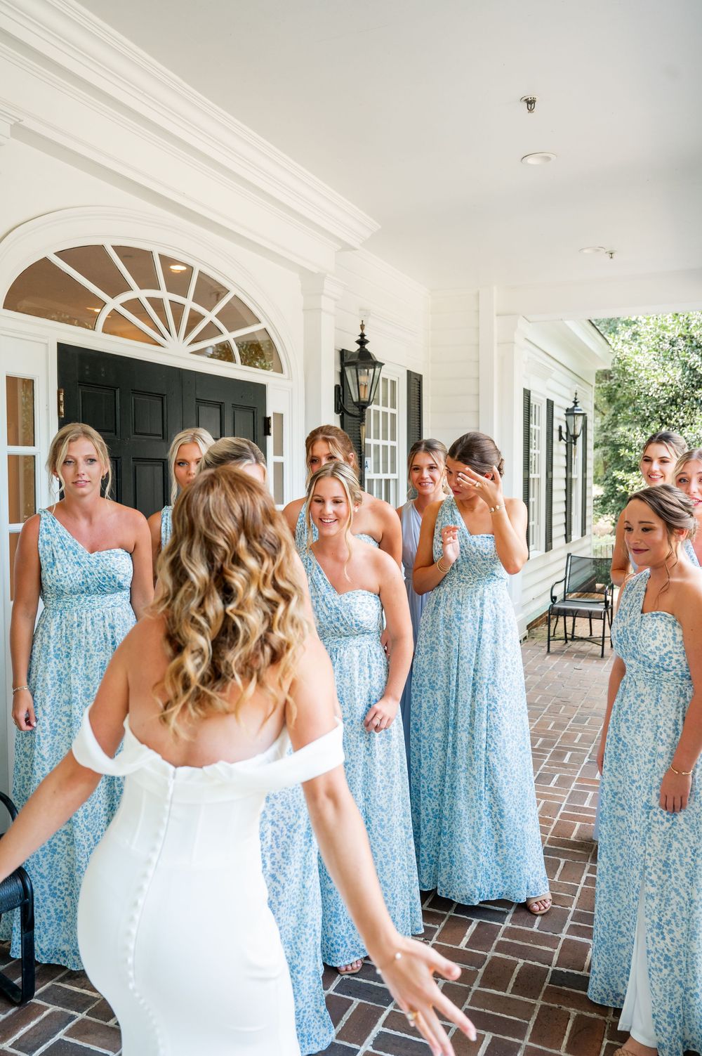 Wedding party wearing coordinated blue dresses stand in a line on a brick porch with white columns.