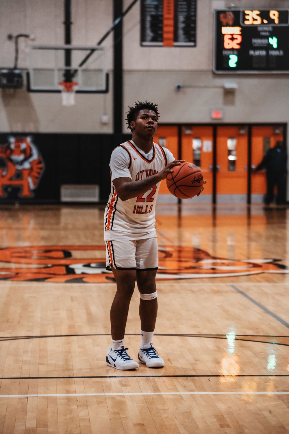 Basketball player prepares to shoot free throw on home court with scoreboard visible.