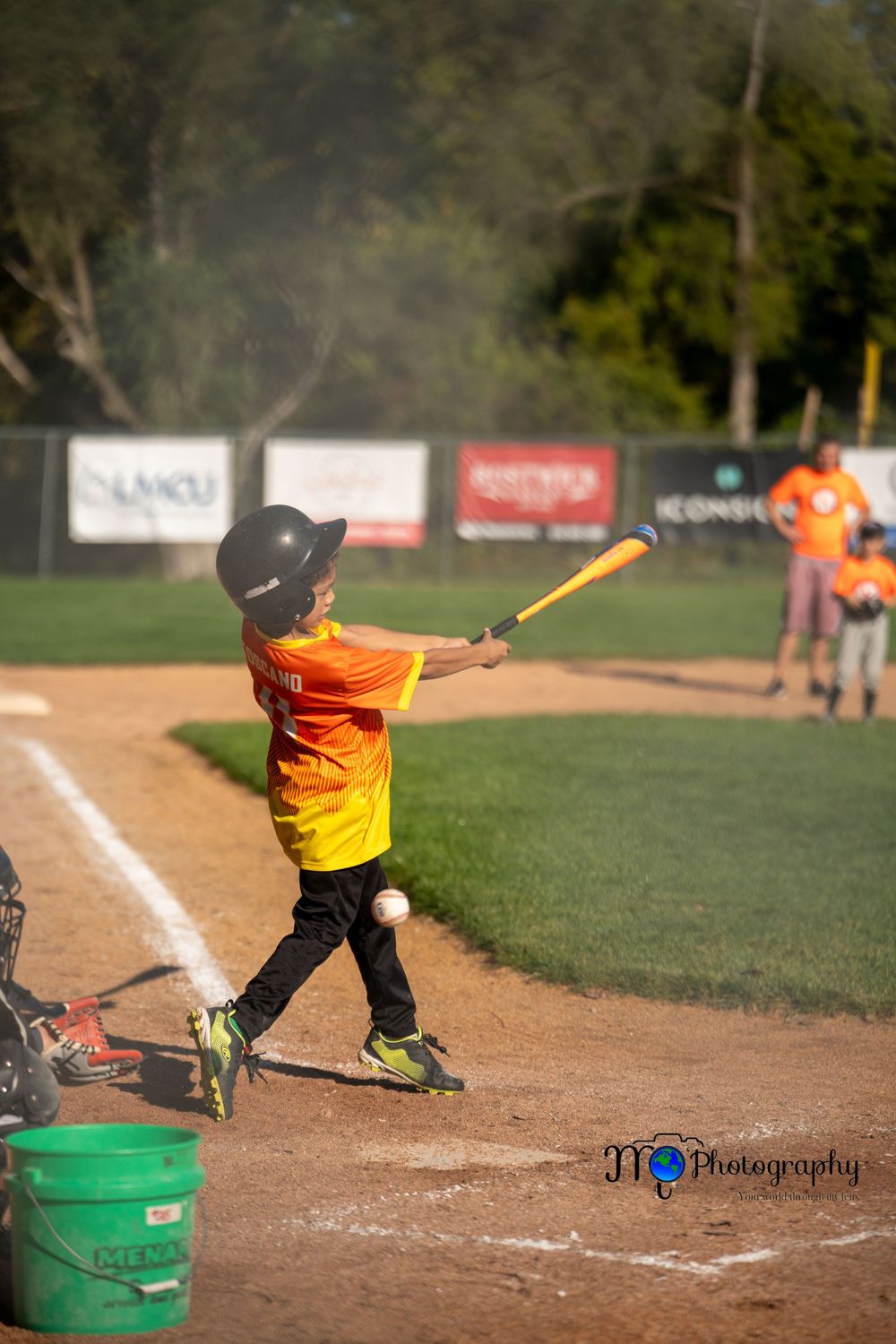 Youth baseball player in orange jersey swings bat at home plate during little league game.