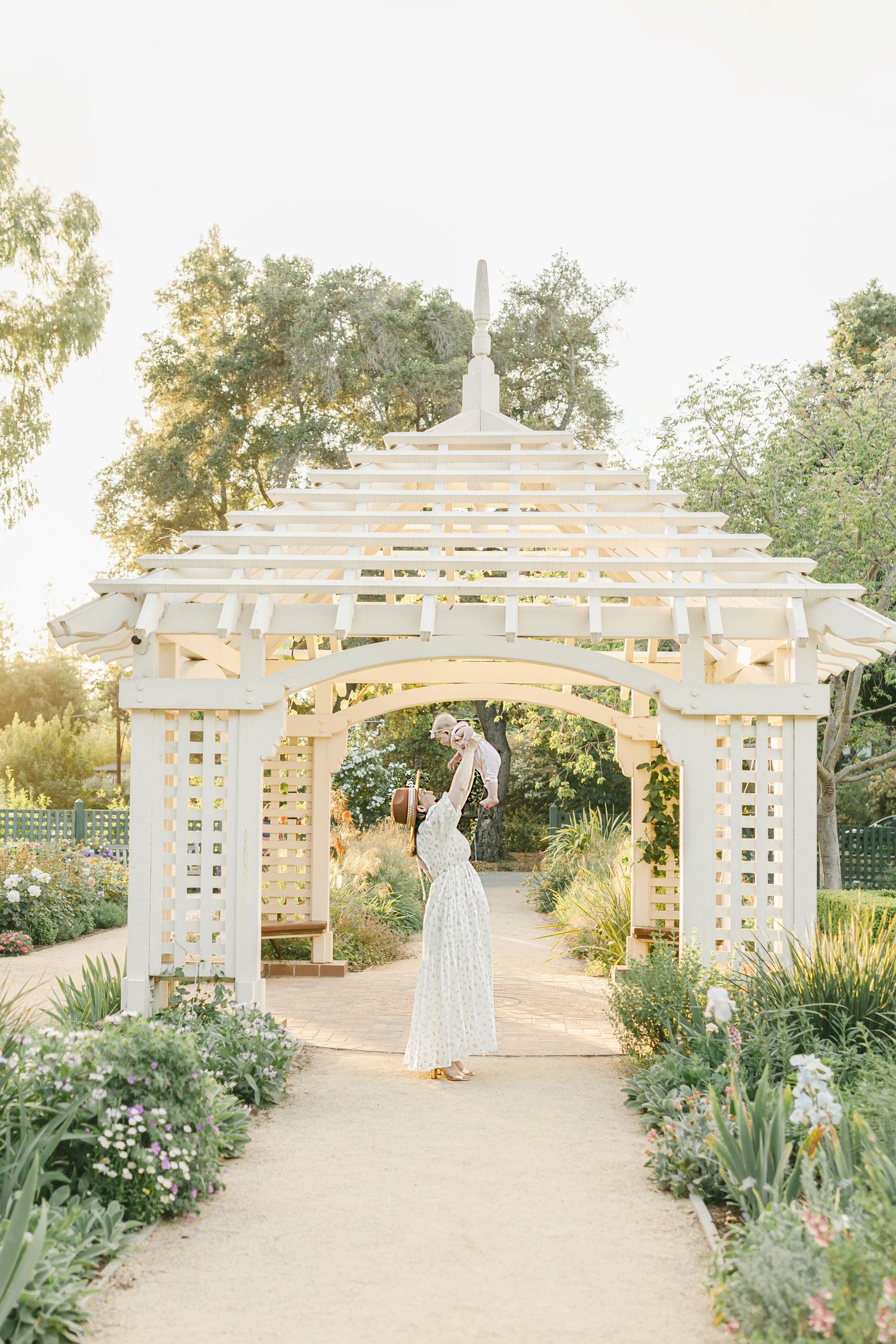 A white wooden garden gazebo with lattice details surrounded by lush landscaping and flowers in soft sunlight.
