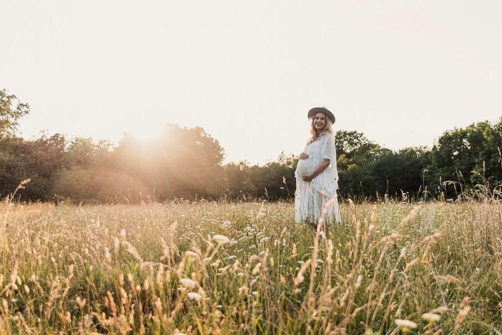 A mother-to-be in white dress stands in golden field of tall grass during sunset with sun flare in background.