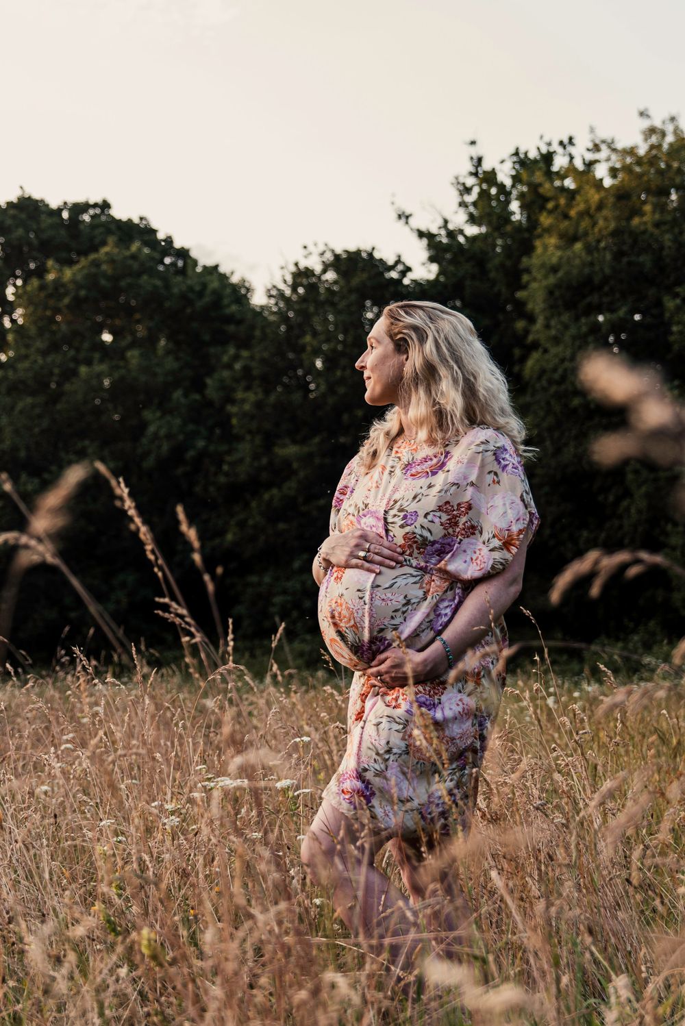 A pregnant woman in a floral dress poses thoughtfully in a golden summer field at sunset.