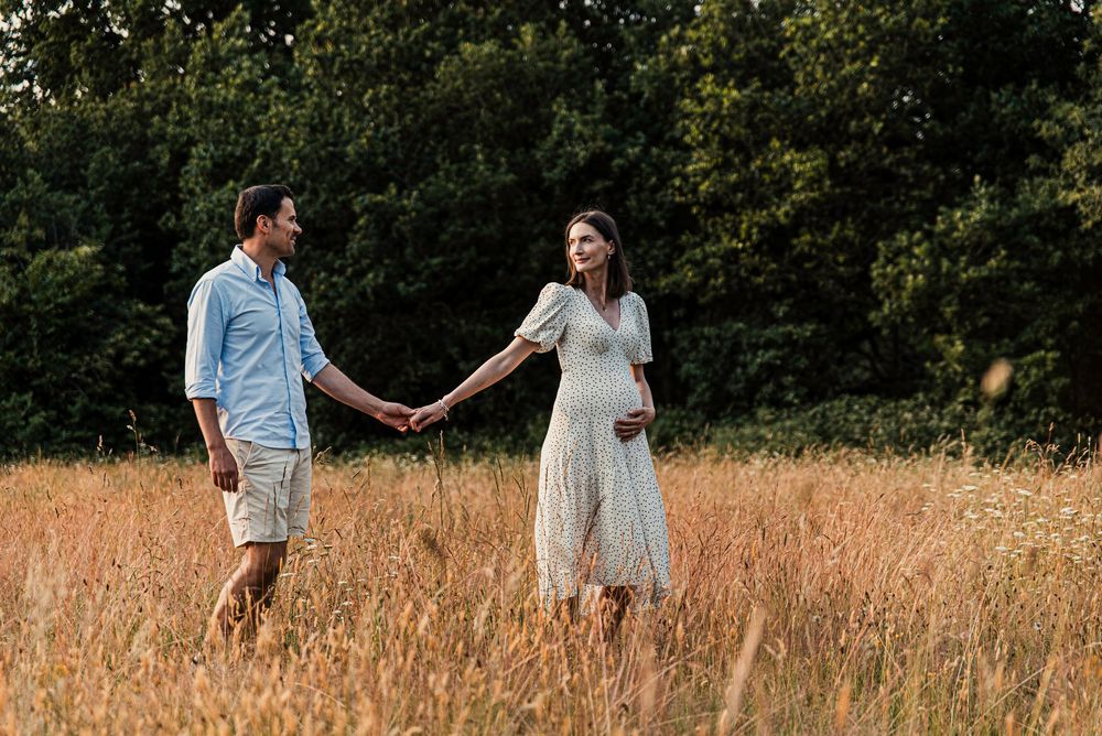 An expectant couple walks hand in hand through a golden wheat field during sunset.