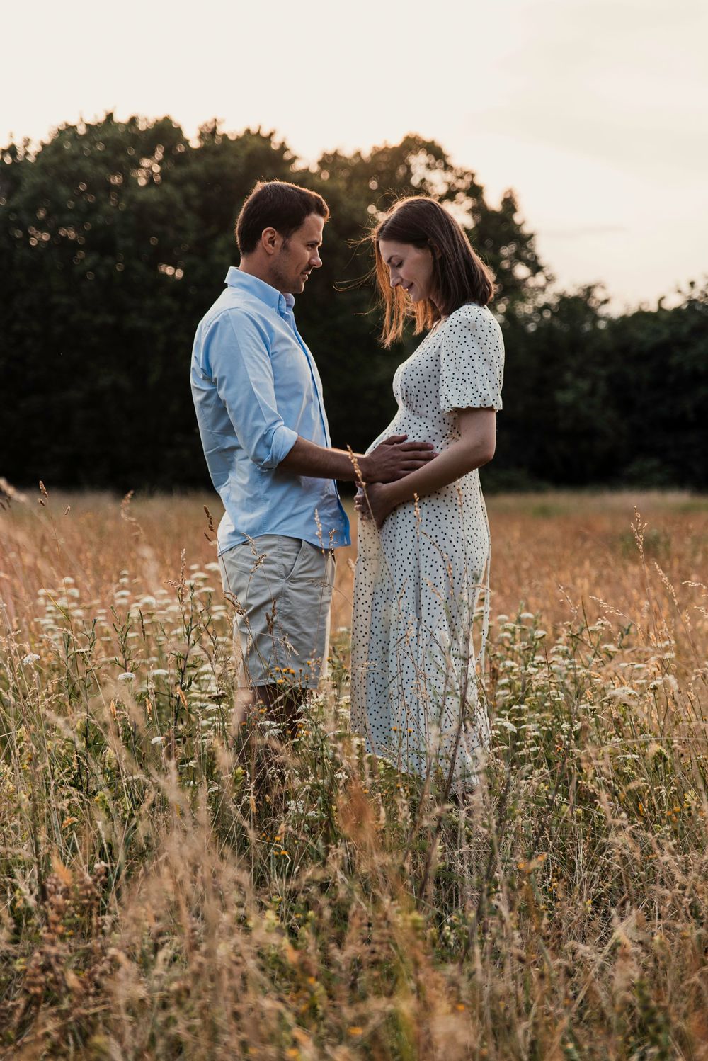 An expectant couple shares a tender moment in a golden field of wildflowers at sunset.