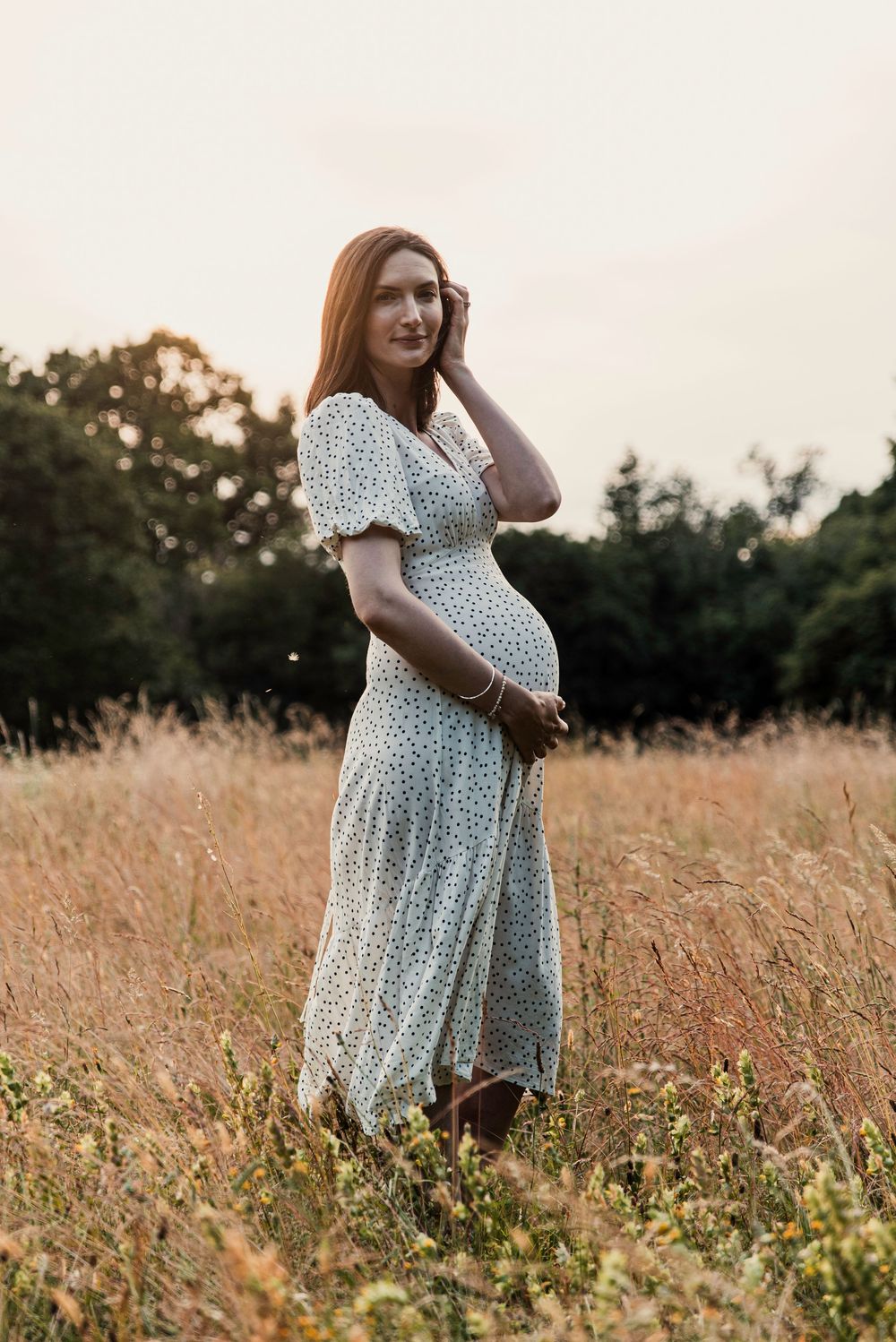 A woman in a polka dot dress stands in a golden field at sunset, cradling her pregnant belly.