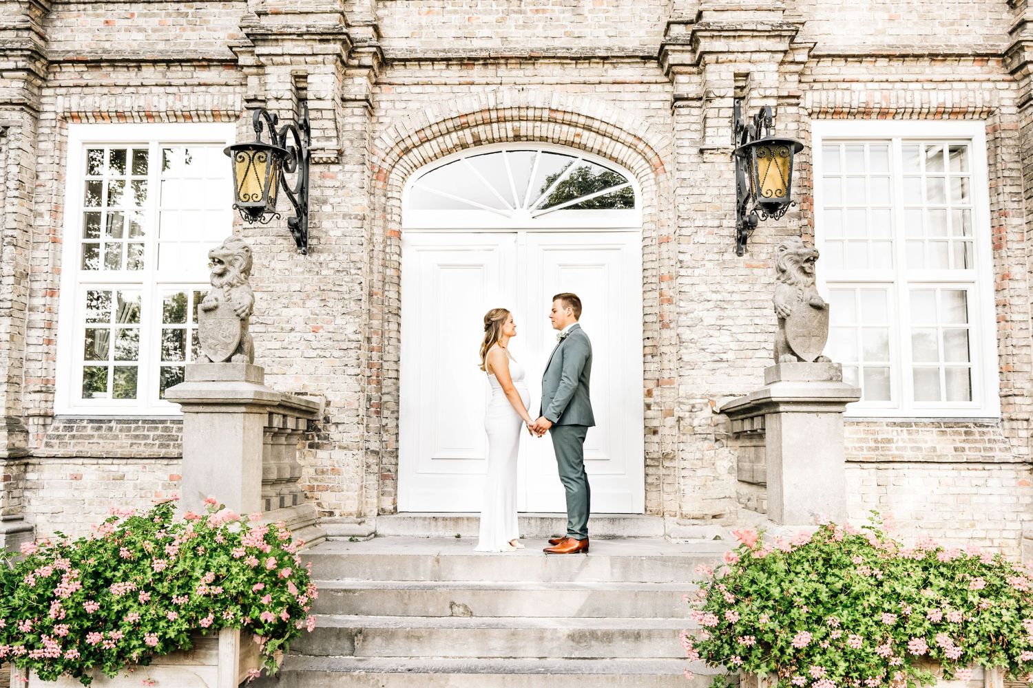 A wedding couple stands elegantly in front of a historic stone building entrance with classic architecture and flowering bushes.
