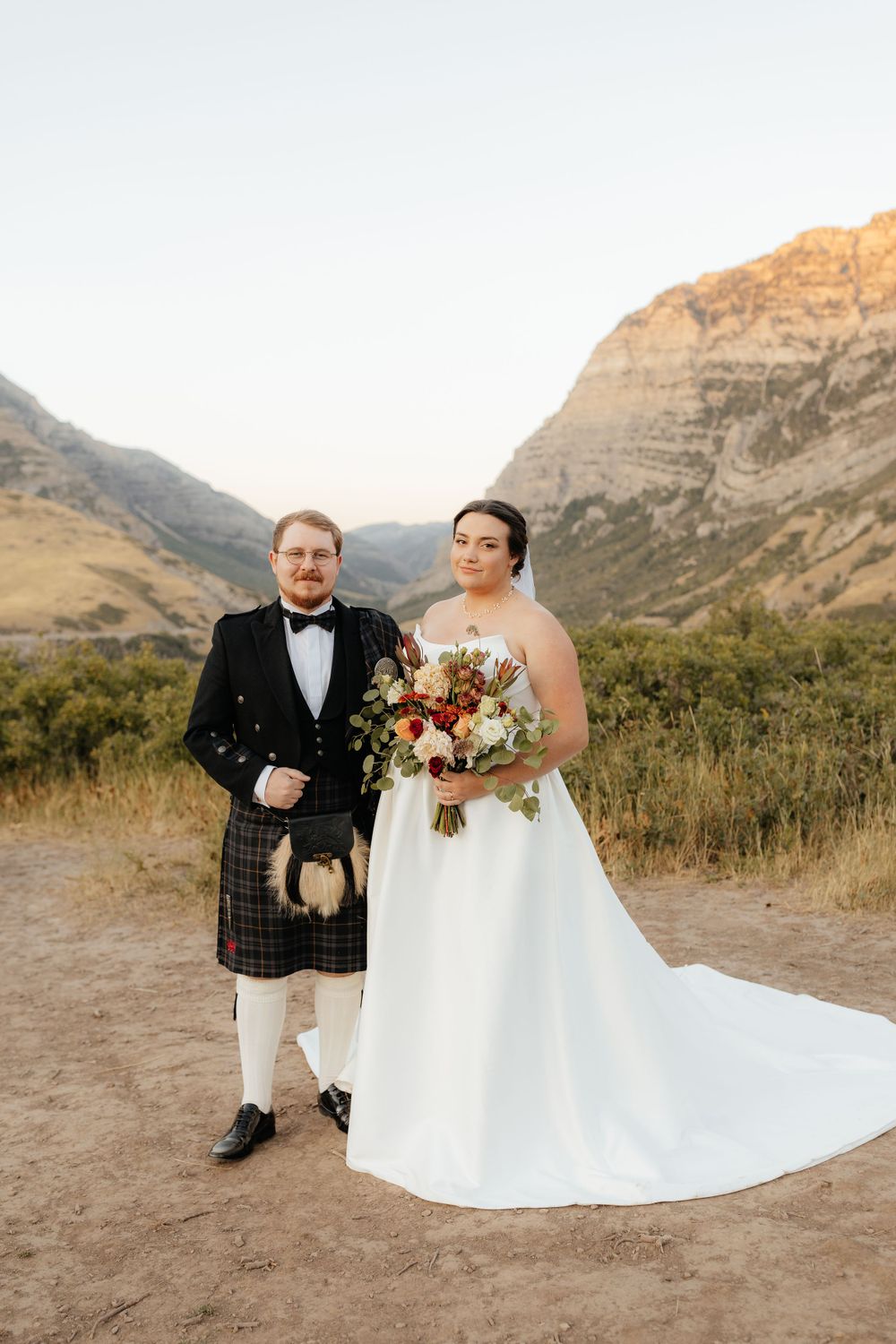 Bride in strapless gown holds autumn bouquet beside groom in Scottish kilt with mountain backdrop.