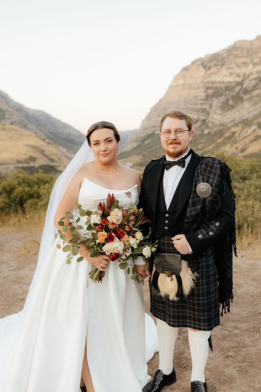 Bride and groom portrait at golden hour, groom in Scottish tartan kilt, mountains in background.