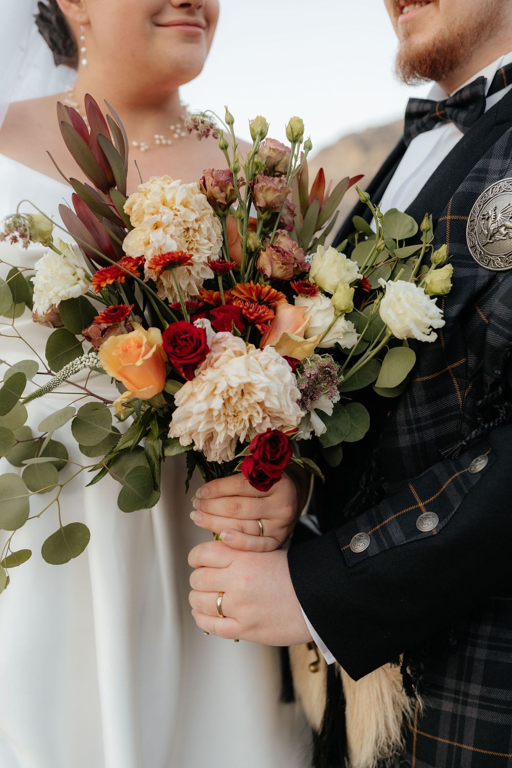 Close-up of bride and groom holding lush bouquet of red, white, and orange flowers together.