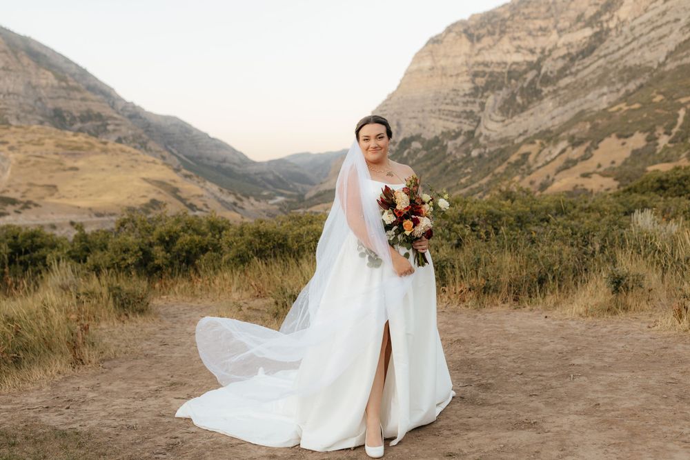 Bride in white gown with long veil poses alone on mountain trail holding rich autumn bouquet.